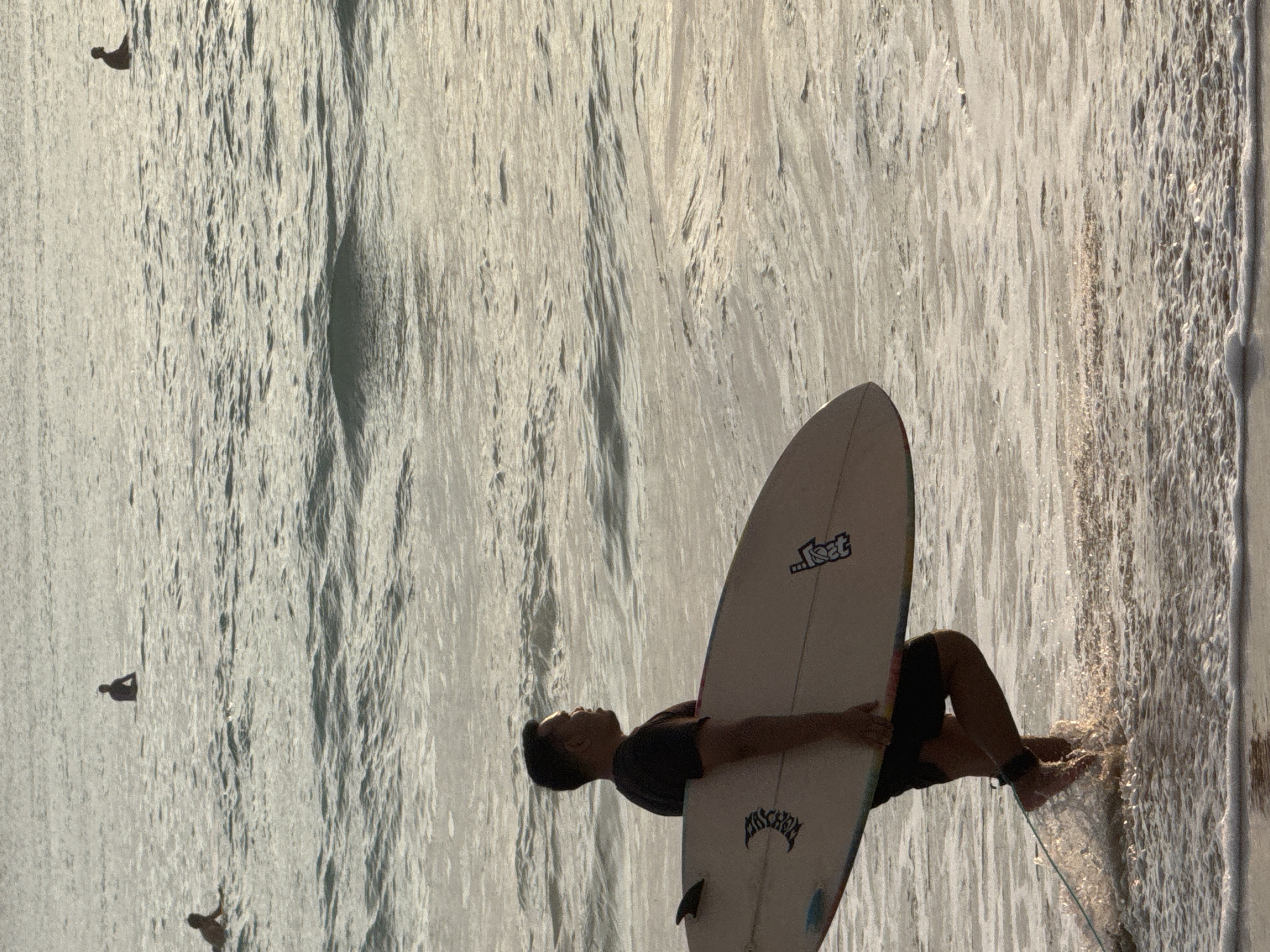 This media file captures a surfer holding a surfboard, lying on his back in shallow ocean water, with waves washing around him. The sun is low in the sky, casting a warm, golden light across the water and creating a shimmering effect on the surface, suggesting it is either early morning or late afternoon. The scene appears to be a beach in Kuta Selatan, Indonesia, given the location context provided. The water is relatively calm, with small, textured waves.
The surfer is silhouetted against the bright water, making his features indistinct, though his dark shirt and swim trunks are visible. He is holding the surfboard vertically, cradling it against his chest. The surfboard is white with visible logos and text. "LOST" is clearly legible on the tail of the board, and above it, there's another logo that appears to be "Lost Surfboards" or similar.
In the background, two other figures, likely surfers or beachgoers, are visible as dark silhouettes in the distance, adding to the sense of a recreational beach scene. The overall mood is tranquil and serene, highlighting the peaceful coexistence of humans and the ocean. The water appears clear, with some sandy patches visible beneath the surface.