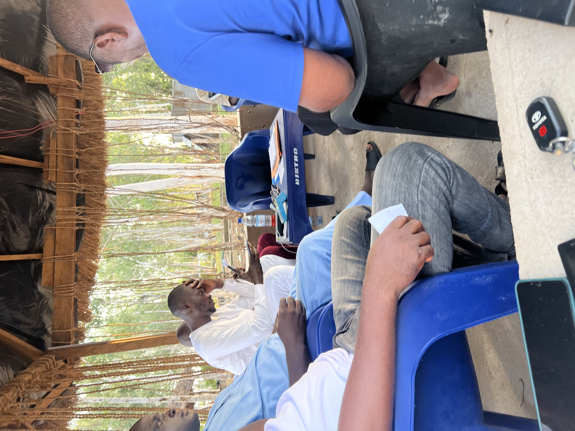 The image shows a group of people gathered outdoors, possibly for a meeting or discussion. The setting appears to be a rustic, open-sided shelter with a thatched roof. The subjects are several men seated on blue plastic chairs around a small, blue plastic table.
One man in the foreground, wearing a white long-sleeved shirt, is sitting with his hand on his forehead, appearing pensive or perhaps uncomfortable. Another man, wearing a bright blue shirt, is also visible, with part of his body obscured by a dark surface, possibly another chair or table. A person in grey pants is seated to the right. A Toyota car key is visible on a textured surface in the lower right quadrant.
On the blue table, there are several objects including a bottle of water, what appears to be a tablet or book, and a stapler or similar office supply. The word "BISTRO" is visible in white text on the side of the blue table.
The scene is lit by natural light, suggesting it is daytime. The background shows greenery and what looks like other structures, possibly indicative of a village or rural area. The overall mood seems informal, with the individuals engaged in their own thoughts or a collective activity. The location cues, such as the rustic shelter and the presence of the Toyota key, are consistent with the provided location context of Mayo Dasa, Nigeria.