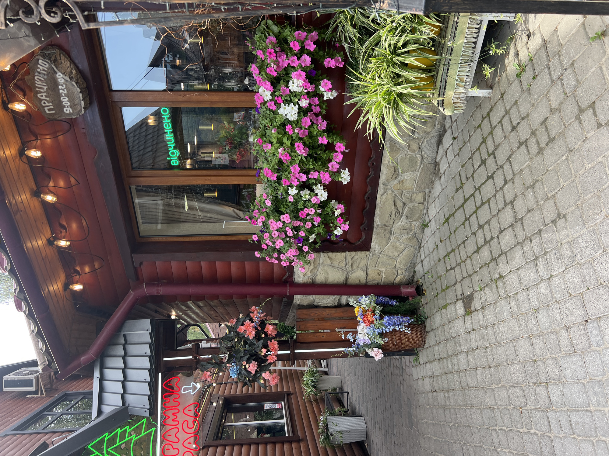 The image shows an outdoor scene in front of a wooden building, likely a cafe or restaurant, in Yaremche, Ukraine. A sign with neon green lettering in Ukrainian that reads "відчинено" (meaning "open") is visible in a window. Above the window, a wooden sign indicates operating hours from 9:00 to 22:00. Red neon signs in Cyrillic characters that appear to say "РАННЯ ПАЦА" are also visible on the left side of the building.
The building features a wooden facade and a roof with visible light bulbs strung along its edge. A red downspout runs along the side of the building. The area in front of the building is paved with cobblestones and decorated with various potted plants and flower arrangements. A large window box filled with pink and white petunias is prominently displayed. In the foreground, a wicker basket holds a bouquet of blue, purple, and orange flowers. Another arrangement of pink flowers hangs in a pot from a wooden post. A small bench covered with a green, fringed textile is visible on the right side of the frame, with several potted plants on it. The overall impression is of a well-decorated and welcoming establishment.