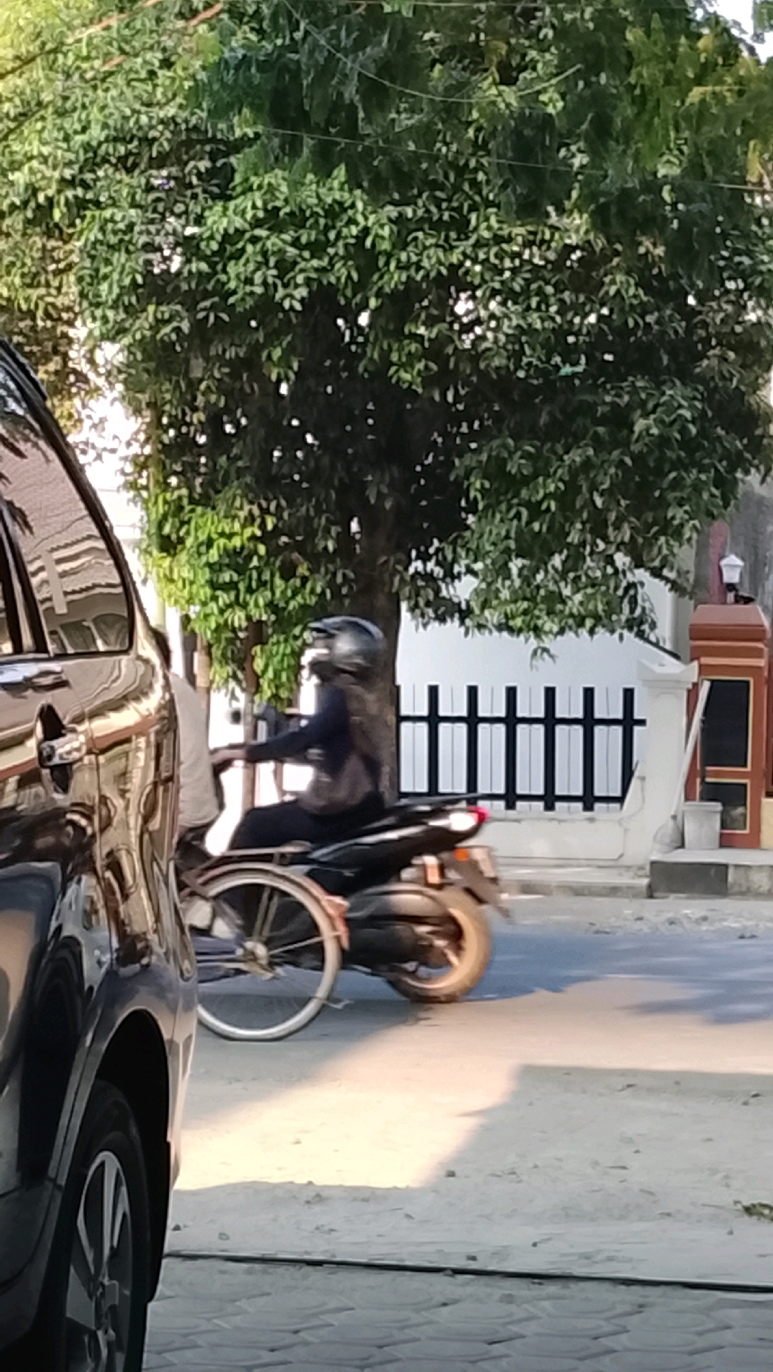 A person wearing a black helmet and dark clothing rides a black motorcycle with a white bicycle attached to the rear, facing left, on a paved street in Slawi, Indonesia. The motorcycle's rear wheel is visible, along with the bicycle's frame and wheel. A dark car is partially visible on the left side of the frame, with its front wheel and side door in view. The background features a lush green tree with dense foliage, a white wall with a black fence, and a brown and grey structure. The lighting suggests it is daytime, with shadows cast on the ground.