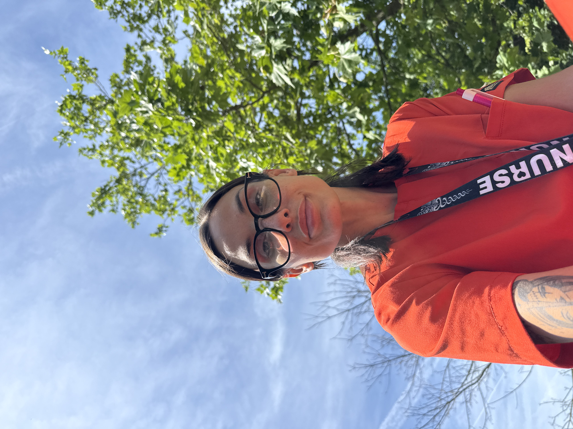 The image captures a selfie of a woman taken from a low angle, looking upwards towards the camera, against an outdoor backdrop in Pleven, Bulgaria. She has dark hair, wears black-rimmed glasses, and displays a calm expression with a slight smile and light-colored lips. Her attire consists of a bright orange or red top, resembling a uniform or scrubs, with a pink and white pen tucked into a pocket on her left sleeve. A prominent black strap with the word "NURSE" in white capital letters and some white patterned symbols is visible across her chest. A tattoo is also visible on her right forearm. The background features a clear, bright blue sky with a few wispy white clouds, indicating a sunny day. Green tree foliage is visible above her, alongside some bare tree branches, suggesting a natural outdoor environment like a park or garden. The lighting indicates it is daytime, likely midday or afternoon.