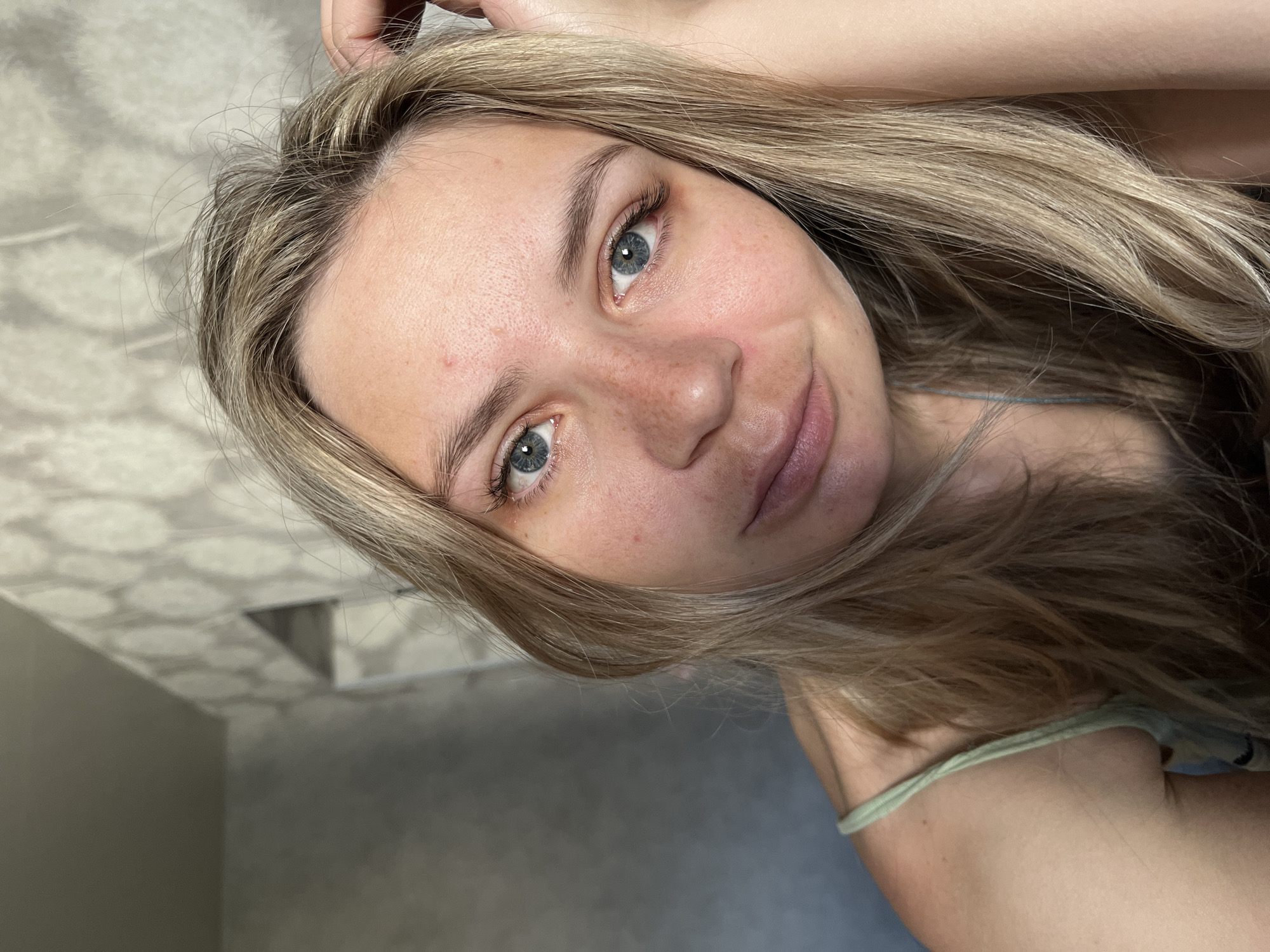 A woman with blonde hair and blue eyes is looking at the camera. She has freckles and slight redness on her face. She is wearing a light green top. The background shows a gray wall and a patterned ceiling. The location is likely Chaikovsky, Russia.