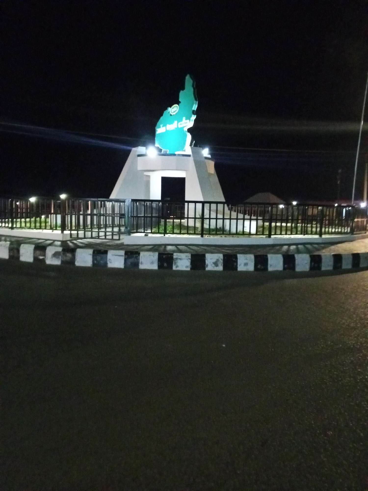 The image depicts a roundabout at night in Unknown, Unknown. A monument is centered in the roundabout, consisting of a white geometric base topped with a green, possibly illuminated, representation of a geographical shape. The base is lit from below. The roundabout is edged with black metal fencing and a black and white painted curb. The surrounding area is dark, with streetlights visible in the background. The foreground shows a dark road surface, possibly wet or reflective. There are no people visible.