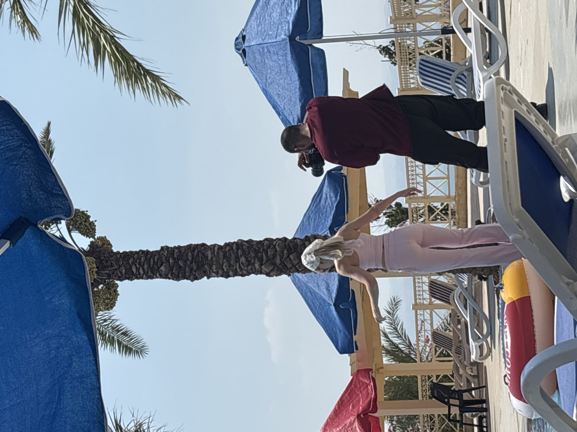 In Ma'in, Jordan, a man in a maroon shirt and dark pants photographs a blonde woman wearing a pink crop top and matching pants, with a white bandana around her head, outdoors near a body of water. The scene is set against a backdrop of palm trees, blue and red umbrellas, and lounge chairs, suggesting a resort or hotel environment. The man holds a camera and is positioned behind the woman, who is facing away from him. The woman appears to be posing or walking. The sky is clear and bright, indicating a sunny day.