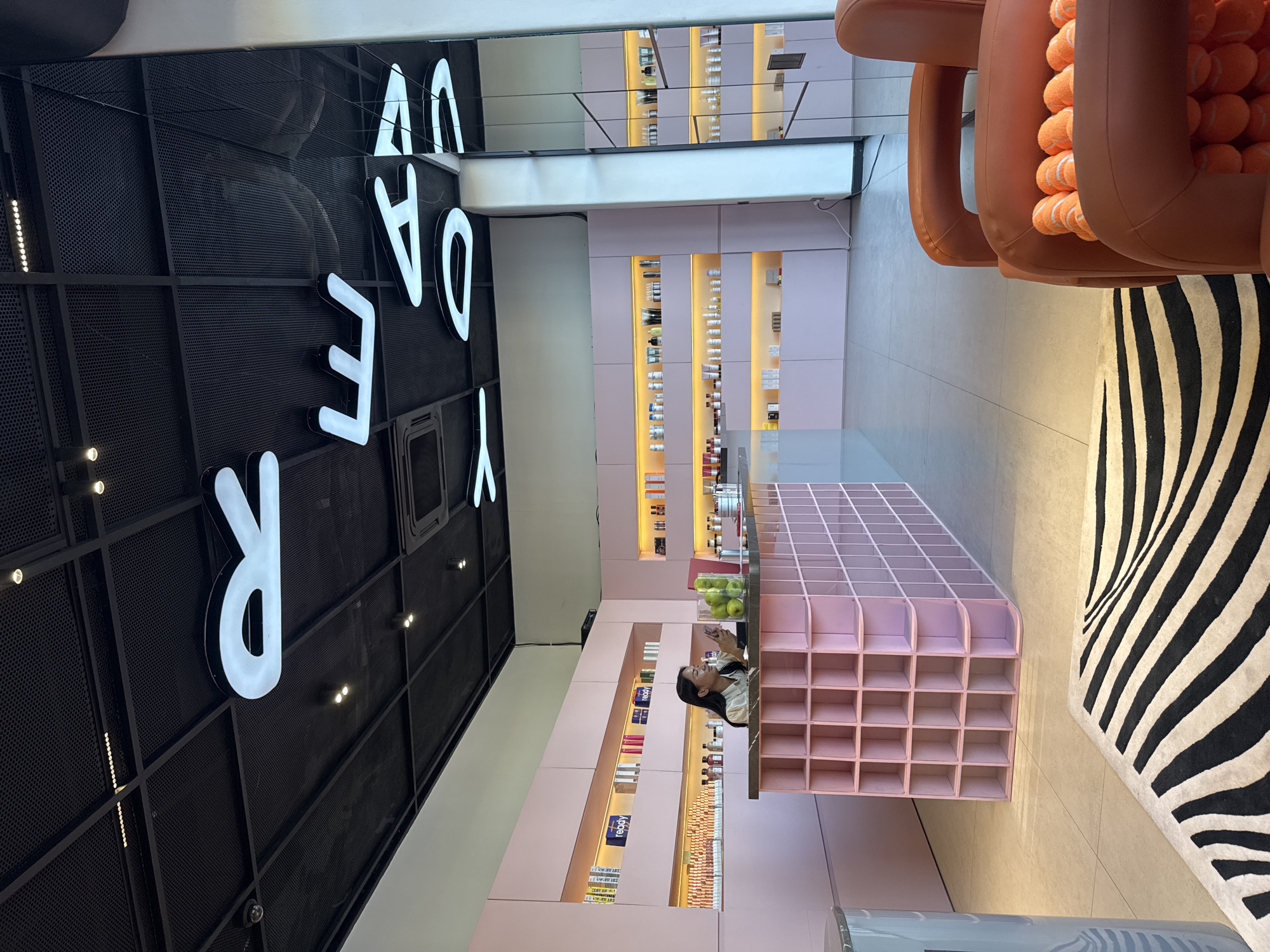The image depicts the interior of a modern retail establishment, possibly a beauty or wellness store, with a distinctly pink and minimalist aesthetic. The space features clean lines and organized displays of products on illuminated shelves lining the walls. A central counter, also pink and segmented into a grid of cubbies, serves as a focal point. A woman with dark hair is visible behind the counter, interacting with what appears to be a mobile device or tablet. In the foreground, a plush, rounded orange armchair is filled with numerous orange tennis balls, creating an unusual decorative element. A zebra-print rug adds a bold contrast to the otherwise pastel environment. Overhead, white, illuminated letters are suspended from the black ceiling, spelling out "READY TO GO," with "READY" partially visible. The store's interior suggests a contemporary and chic ambiance. The presence of numerous skincare or cosmetic products on display implies a retail environment specializing in such items. The overall impression is one of a stylish and welcoming space designed for shopping and possibly consultation. This establishment is located in Kuta Selatan, Indonesia.