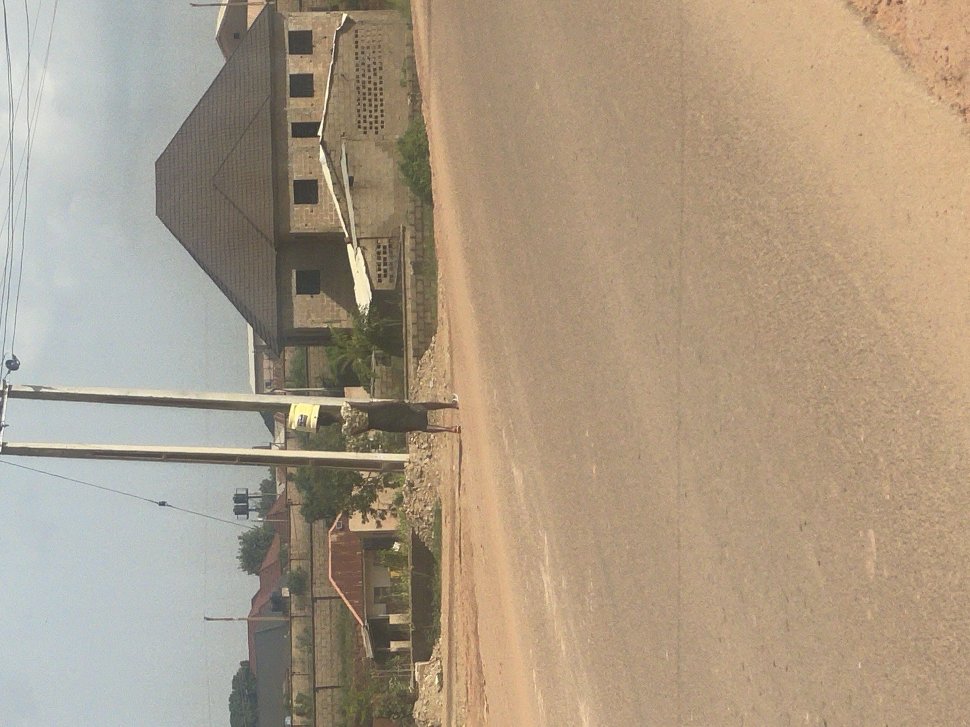 A woman is walking on a road in Kwang, Nigeria, carrying a yellow bucket on her head. She is wearing a patterned top and a dark skirt. In the background, there are several buildings, including a house with a brown tiled roof and a smaller structure with a corrugated metal roof. Power lines and utility poles are visible in the scene, suggesting an urban or semi-urban environment. The road appears to be unpaved, with dirt and gravel on the sides. The sky is clear and the lighting suggests it is daytime.