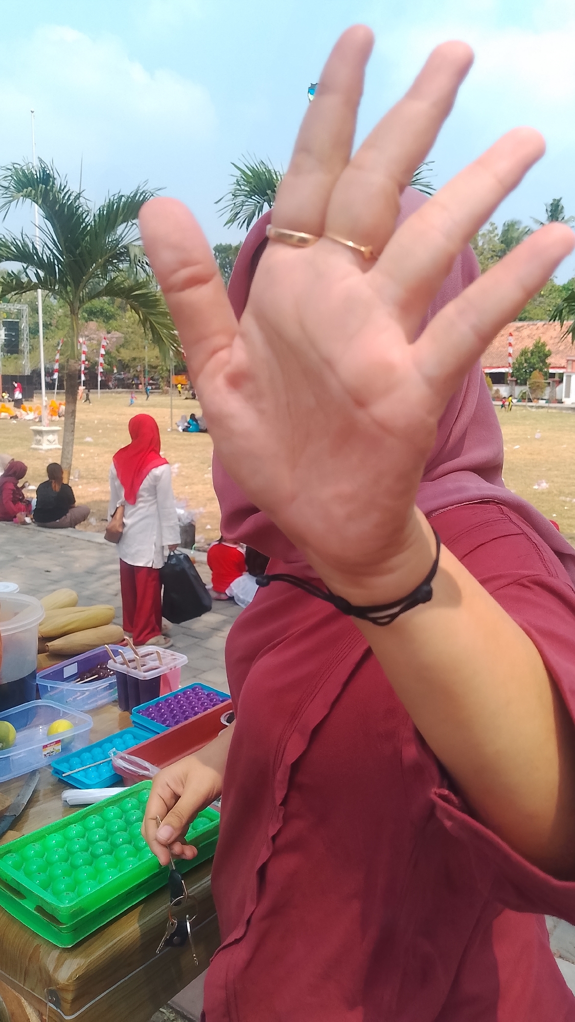 In the city of Karanglangu, Indonesia, on a sunny day, a person wearing a maroon headscarf and shirt is holding their hand up to the camera, obscuring their face. They are wearing two gold rings on their left hand and a black bracelet on their wrist. In the foreground, a wooden table displays various containers of colorful items, including ice cube trays filled with green spherical objects, purple spheres, and what appear to be popsicles. A knife rests on the table. In the background, people are seated on a grassy field, and palm trees line the area. The scene appears to be an outdoor gathering or event.
