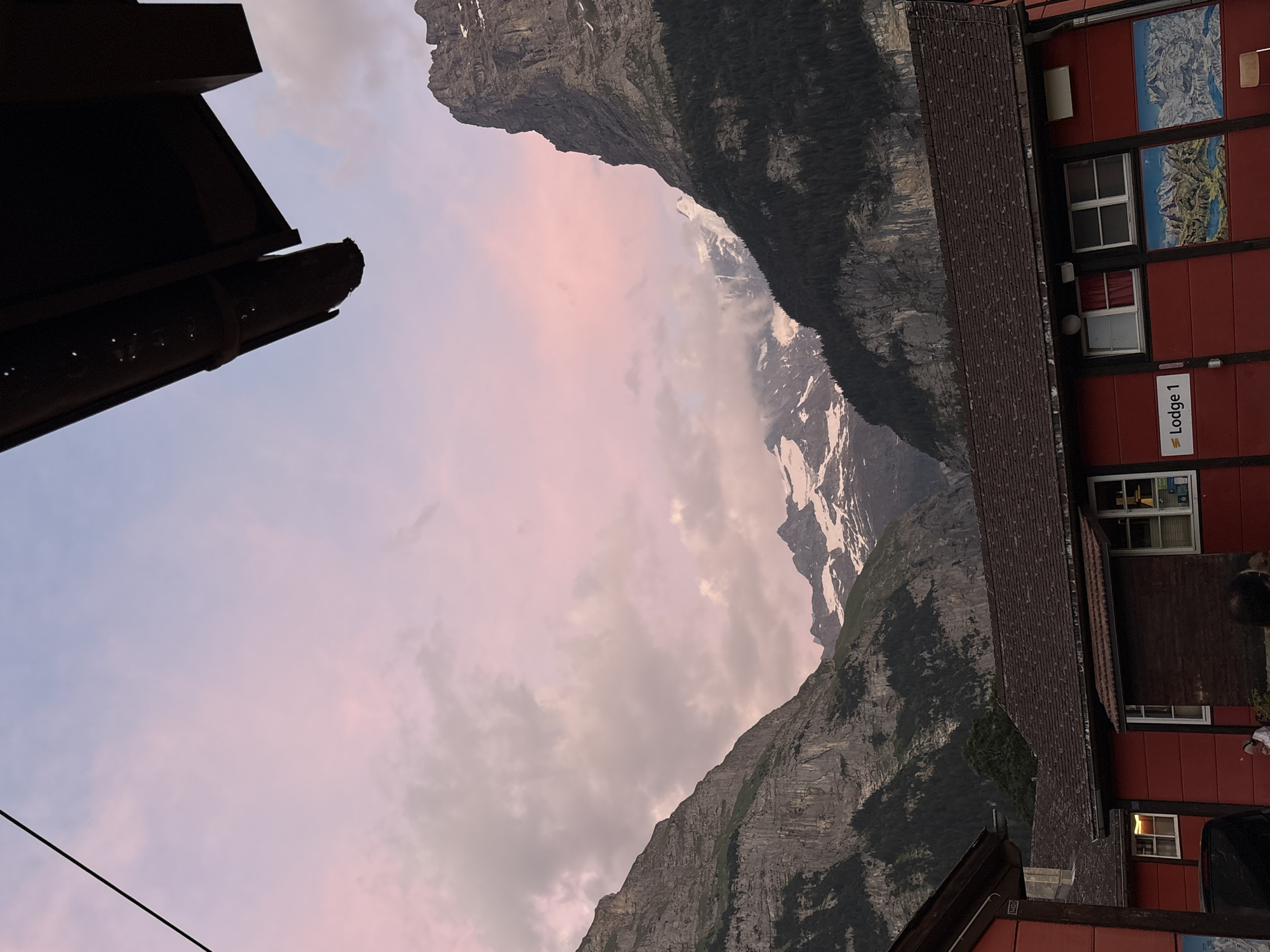 The image depicts a view of the Swiss Alps from a building in Grindelwald, Switzerland. In the foreground, a red building with a dark tiled roof is visible. The building has several windows and two framed images of mountain landscapes mounted on its facade. One image is labeled "Lodge 1". Above the building, the rugged slopes of the mountains rise sharply, covered in patches of green vegetation and dark green pine trees. Snow-capped peaks are visible in the distance, partially obscured by clouds. The sky above is a soft gradient of pink and blue, suggesting either sunrise or sunset. The overall scene conveys a sense of natural beauty and tranquility, with a hint of human presence from the building and the people partially visible in front of it.