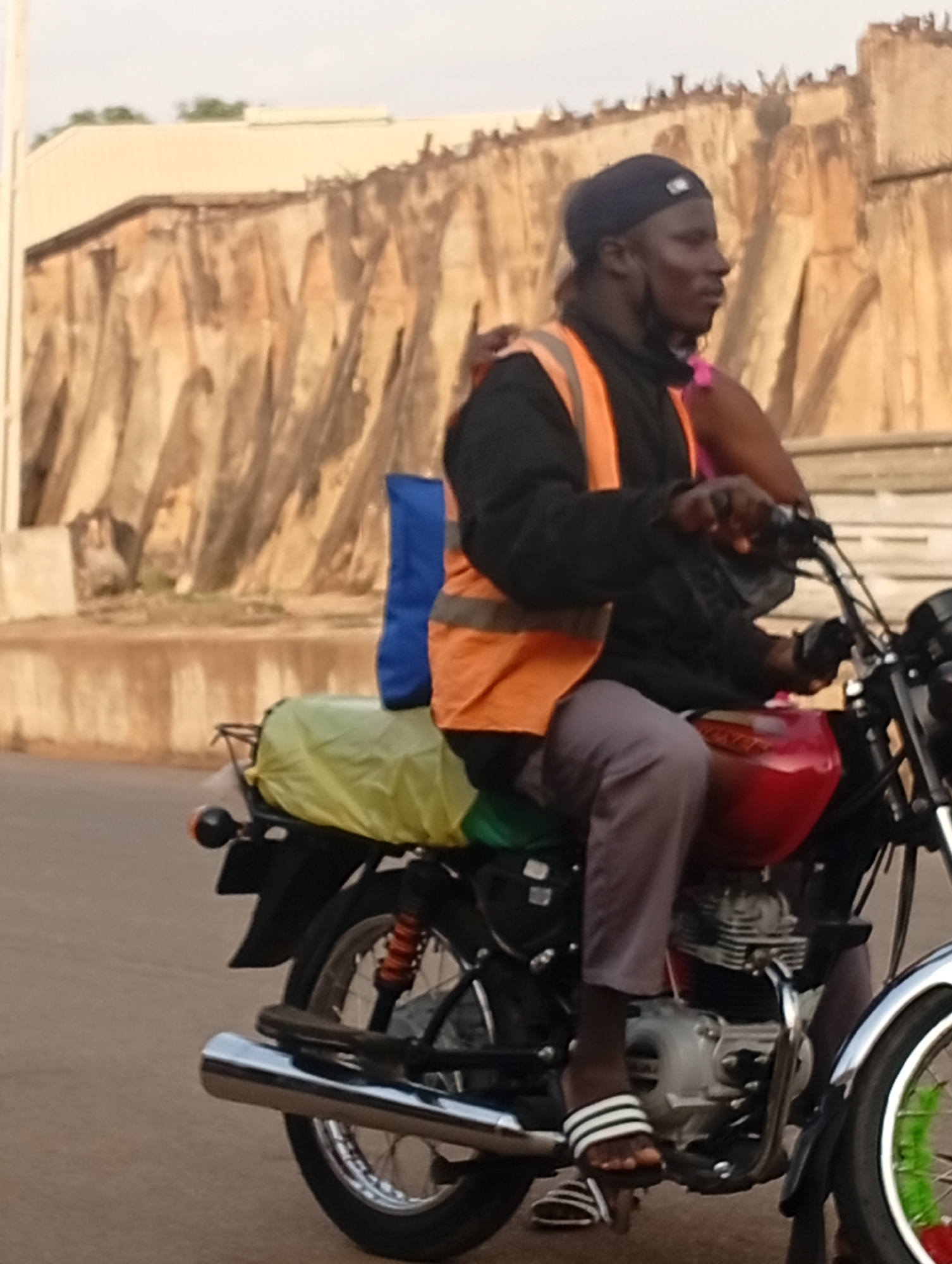 A man is riding a red motorcycle on a paved road in Gura Top, Nigeria. He is wearing a black beanie, a black shirt, an orange safety vest, grey pants, and black and white striped sandals. A passenger is behind him. A blue bag and yellow sheet is on the motorcycle carrier. The background includes a beige-colored wall.