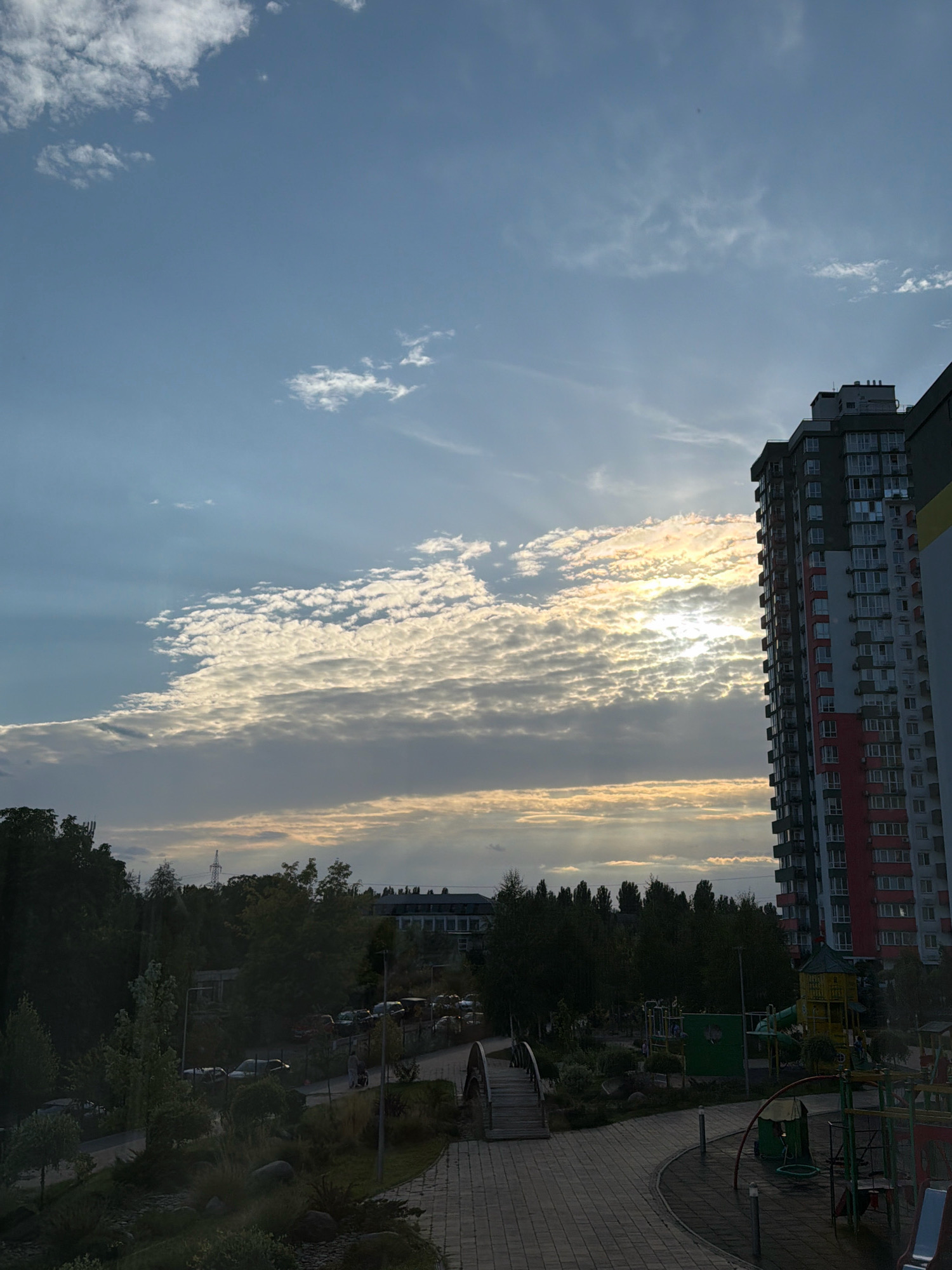 The image depicts an outdoor scene in a residential area of Kyiv, Ukraine, likely during late afternoon or early evening, given the quality of light. Dominating the right side of the frame is a multi-story apartment building with a modern facade, featuring a mix of gray, white, and red sections, with numerous windows. To the left of the building, a band of clouds stretches across the sky, catching the sunlight and creating a bright, textured atmospheric effect. The sky itself is a clear blue, with scattered smaller clouds.
Below the apartment building and the sky, a verdant landscape unfolds. Trees line the horizon, forming a dense green buffer. In the foreground, a playground area is visible, with various colorful structures, including slides and swings, set on a paved surface. A small, arched pedestrian bridge is also present, crossing over a landscaped area. A paved walkway curves through the scene. In the background, beyond the trees, there appear to be other buildings and infrastructure, including what looks like a power pylon. Several parked cars are visible along a road. The overall impression is one of a well-developed urban residential space with recreational amenities.