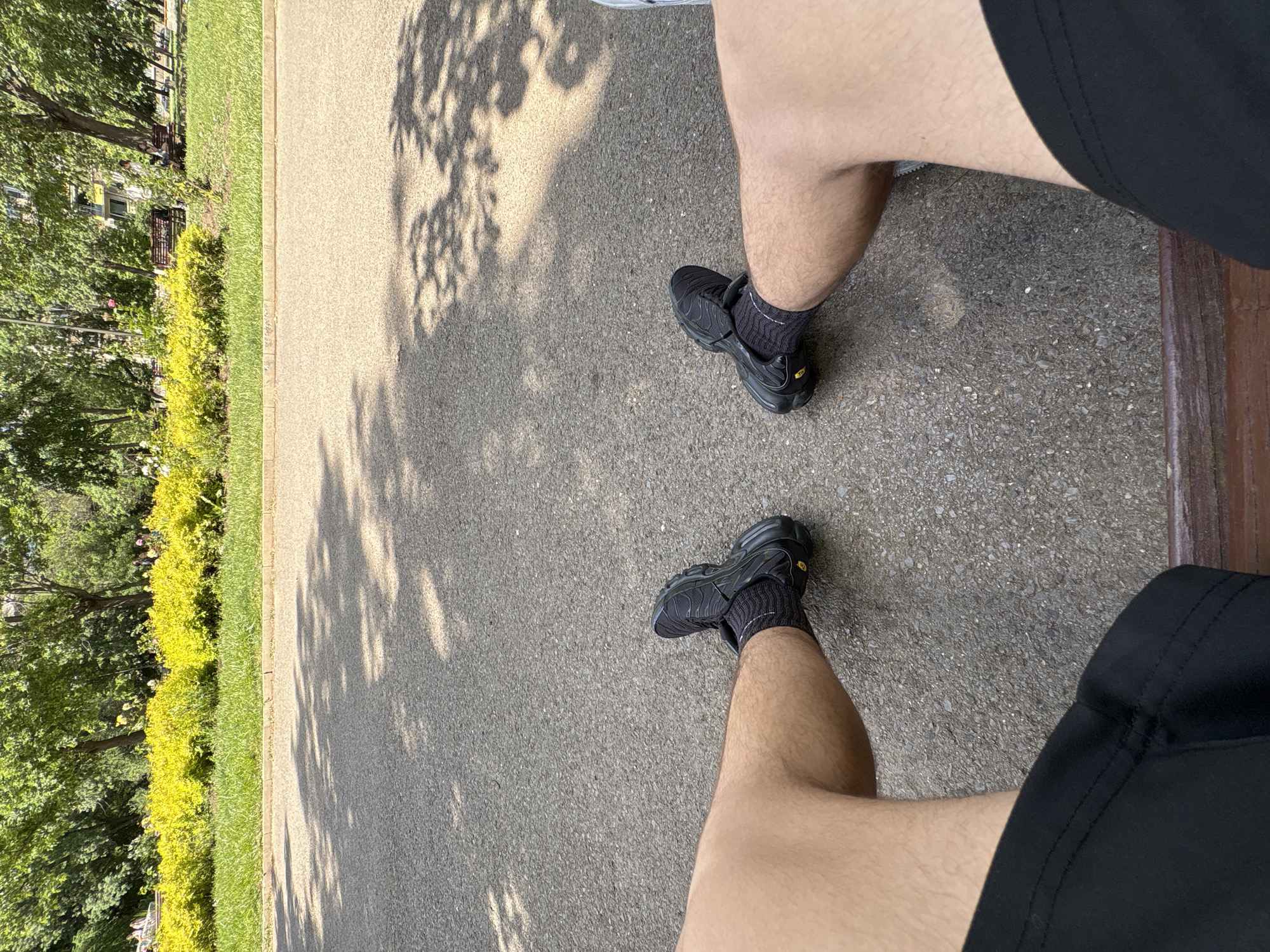 The image shows a first-person view of two legs in shorts and athletic shoes standing on a paved surface. The legs are positioned on the right side of the frame. To the left, there is a strip of green grass and bushes, followed by trees. Shadows of leaves are cast on the paved surface. A portion of a wooden bench is visible on the right side of the frame.