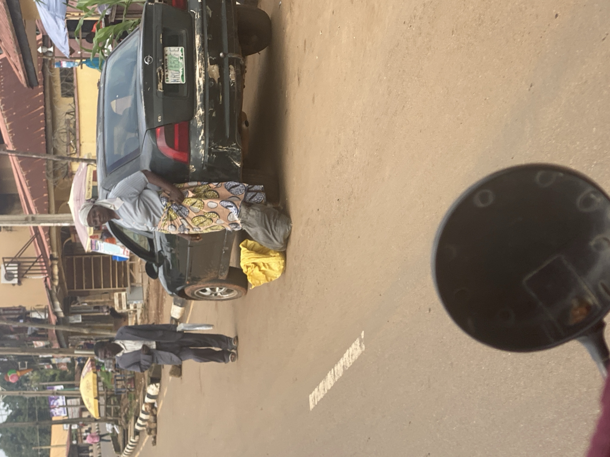 The image depicts a street scene in Maigizo, Nigeria. In the foreground, the reflection of a black motorcycle's mirror is visible. To the left, a dark-colored sedan is parked, its rear end facing the viewer. A woman, dressed in a light-colored shirt and a patterned skirt, is leaning against the car, holding a white cloth over her shoulder. Next to her, a yellow bag rests on the ground. Further down the road, a man in a dark suit and white shirt walks away from the camera. The street is paved with asphalt and has a white painted line marking. Buildings with corrugated iron roofs and balconies line the street in the background. The scene appears to be set during daytime, with no clear indications of weather conditions. The overall impression is one of everyday life on a street in a Nigerian town. A license plate on the car reads "HRD 552 FE".