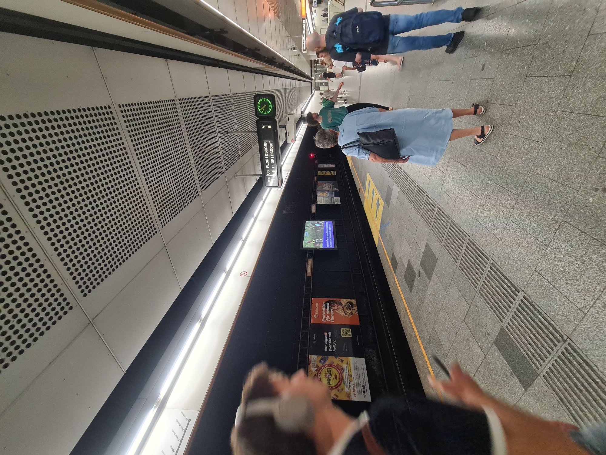 The image shows a view of a subway station platform. In the foreground, a person wearing headphones is partially visible on the left, looking towards the right. On the platform, several people are waiting, some are walking. A digital display board indicates "U6 FLORIDSDORF" and a clock is visible. Posters are displayed on the wall behind the platform. The platform itself is paved with tiles and has tactile paving for the visually impaired. The ceiling features a grid of lights and perforated panels. The overall environment appears to be a modern subway station.