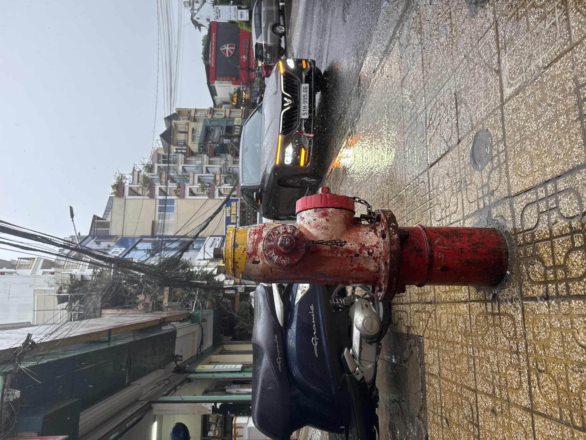 The image captures a street scene in Da Lat, Vietnam, during a rainy day. A red and yellow fire hydrant stands prominently in the foreground, slightly obscuring a parked blue Yamaha Grande scooter. Behind these objects is a black VinFast car with license plate "51H-995.66".
The background features a multi-story building complex across the street, with a restaurant displaying an emblem resembling a red cross on a white circle. Overhead, a dense network of tangled electrical wires fills the sky. The street is wet, reflecting the lights of the car and the surrounding buildings. The tiled sidewalk is also wet.