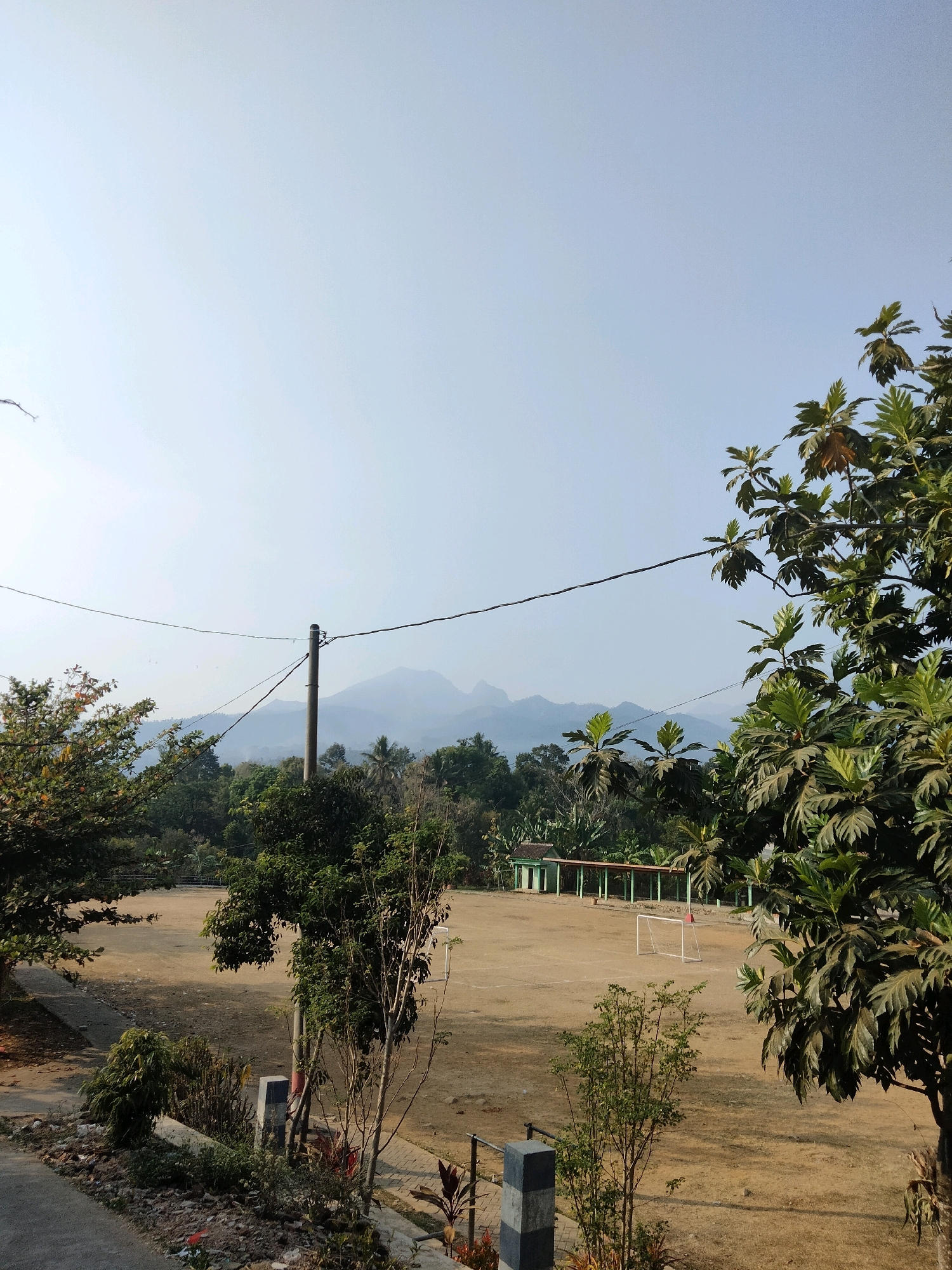 This is a medium shot, outdoor, daytime photograph taken in Sawahan, Indonesia. The scene captures a rural landscape with a dirt sports field in the foreground. A soccer goal is visible on the field, along with a covered structure or bleachers in the background. Lush green trees and vegetation surround the field. In the distance, hazy mountains are visible under a clear sky. A utility pole with wires is positioned in the middle ground, adding to the rural atmosphere. The lighting suggests it is a sunny day.