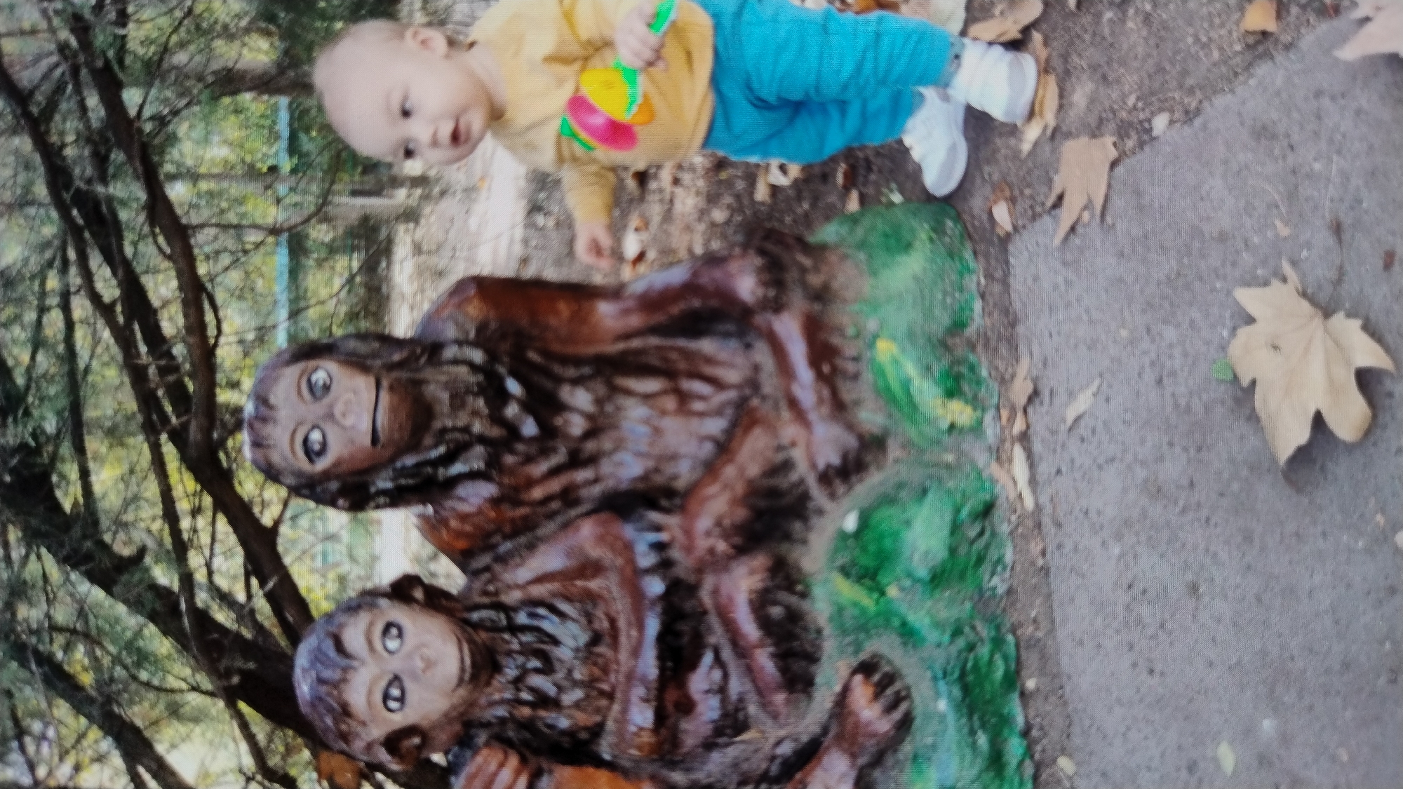 The image depicts a young child, possibly a toddler, standing to the right of two large, dark brown, sculpted figures that resemble primates. The figures are positioned side-by-side and are seated on a green, textured base. The child is facing away from the camera, looking towards the primate sculptures. They are wearing a yellow long-sleeved shirt with a colorful, circular design on the front, bright blue pants, and white shoes. The child is holding a green object with a colorful, multi-faceted tip.
The sculptures and the child are situated outdoors, with a natural environment visible in the background. Trees with branches and foliage form the backdrop, suggesting a park or a wooded area. The ground in front of the sculptures appears to be a paved path, with fallen leaves scattered across it. The overall scene suggests a casual, outdoor setting where the child is observing or interacting with the sculptures.
The image does not provide any specific geographical markers or elements that would allow for the identification of the city of Mykolaiv, Ukraine.