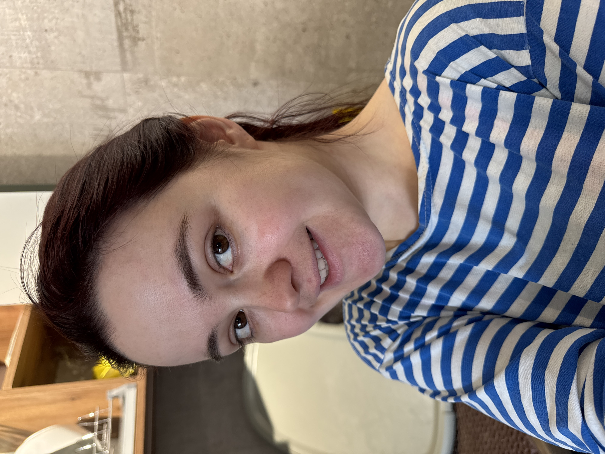 A young woman is depicted in a close-up portrait, taken indoors. The image is oriented sideways, with her head tilted towards the right. She has dark brown hair, dark brown eyes, and fair skin with a natural, light blush and faint freckles across her nose and cheeks. She looks towards the viewer with a gentle, friendly smile, revealing her upper teeth. She is wearing a casual long-sleeved top with distinctive horizontal blue and white stripes.
The background suggests a casual home environment. Behind her head and to the upper left, a light-colored wall or tiled surface is visible. To her lower left, part of a wooden cabinet or shelf can be seen, along with some metallic, reflective objects, possibly a dish rack. To her right and below her, a darker, possibly carpeted or mat area is present. The lighting is soft and even, illuminating her face clearly. The woman's expression is calm and pleasant. There is no visible text in the image. The context indicates the location is Izhevsk, Russia.