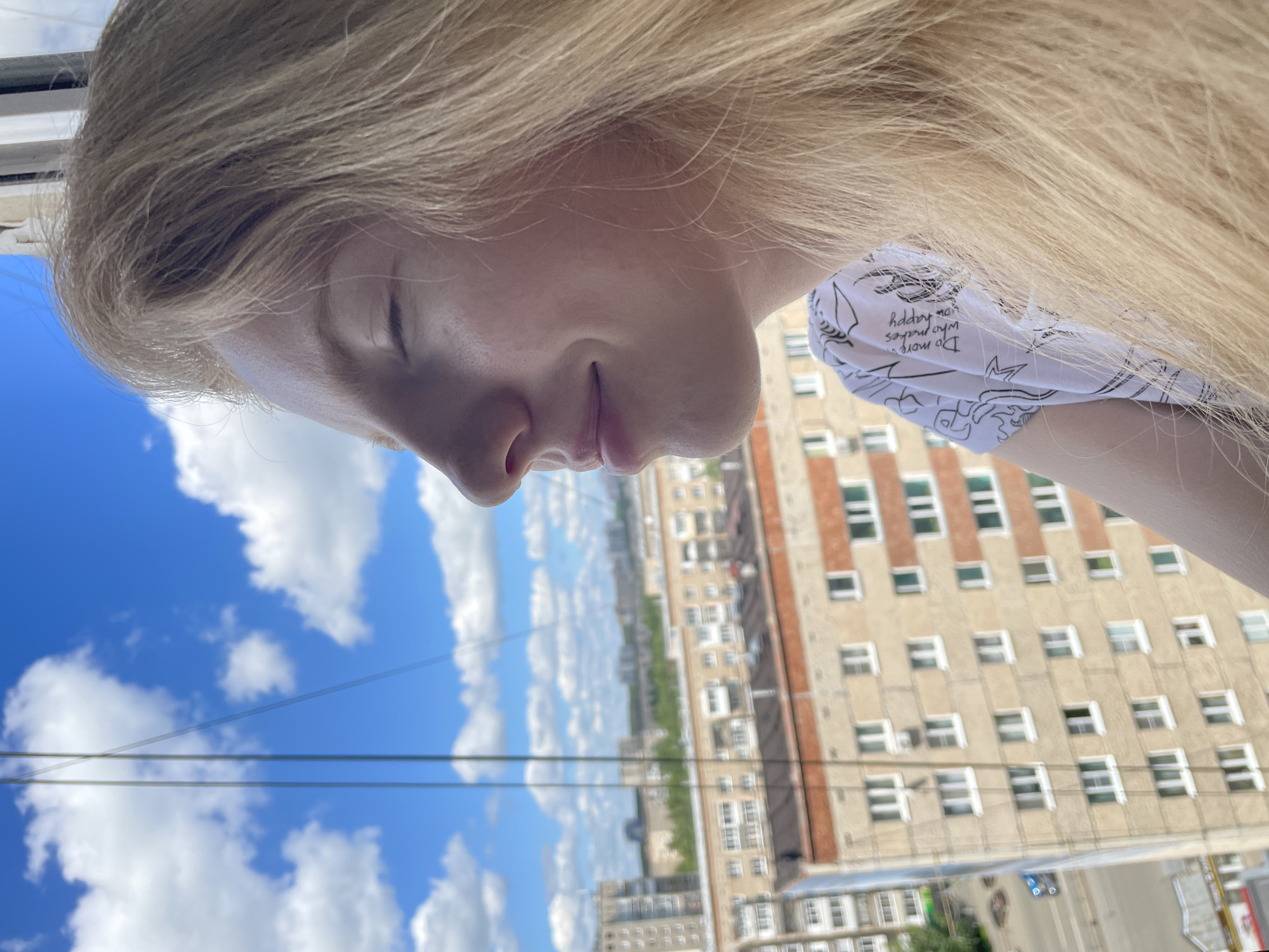 The image, rotated 90 degrees clockwise, presents a close-up profile of a person with long, flowing blonde hair. The person's eyes are closed or cast downwards, and their lips are slightly parted, conveying a serene or contemplative expression. They are wearing a white garment adorned with black text and abstract drawings. Partially visible text on the garment reads "Do more who makes you happy."
The background features a vibrant blue sky filled with scattered white cumulus clouds, indicating a bright, sunny day. Two thin, dark lines, likely utility wires, horizontally traverse the sky. Below the sky, a distant urban landscape is visible, comprising multiple light-colored, multi-story buildings with numerous rectangular windows. The architectural style suggests a city environment, consistent with Izhevsk, Russia. The person appears to be positioned at an elevated point, possibly a window or balcony, overlooking the city. The time of day is clearly daytime, with strong natural light illuminating the scene.