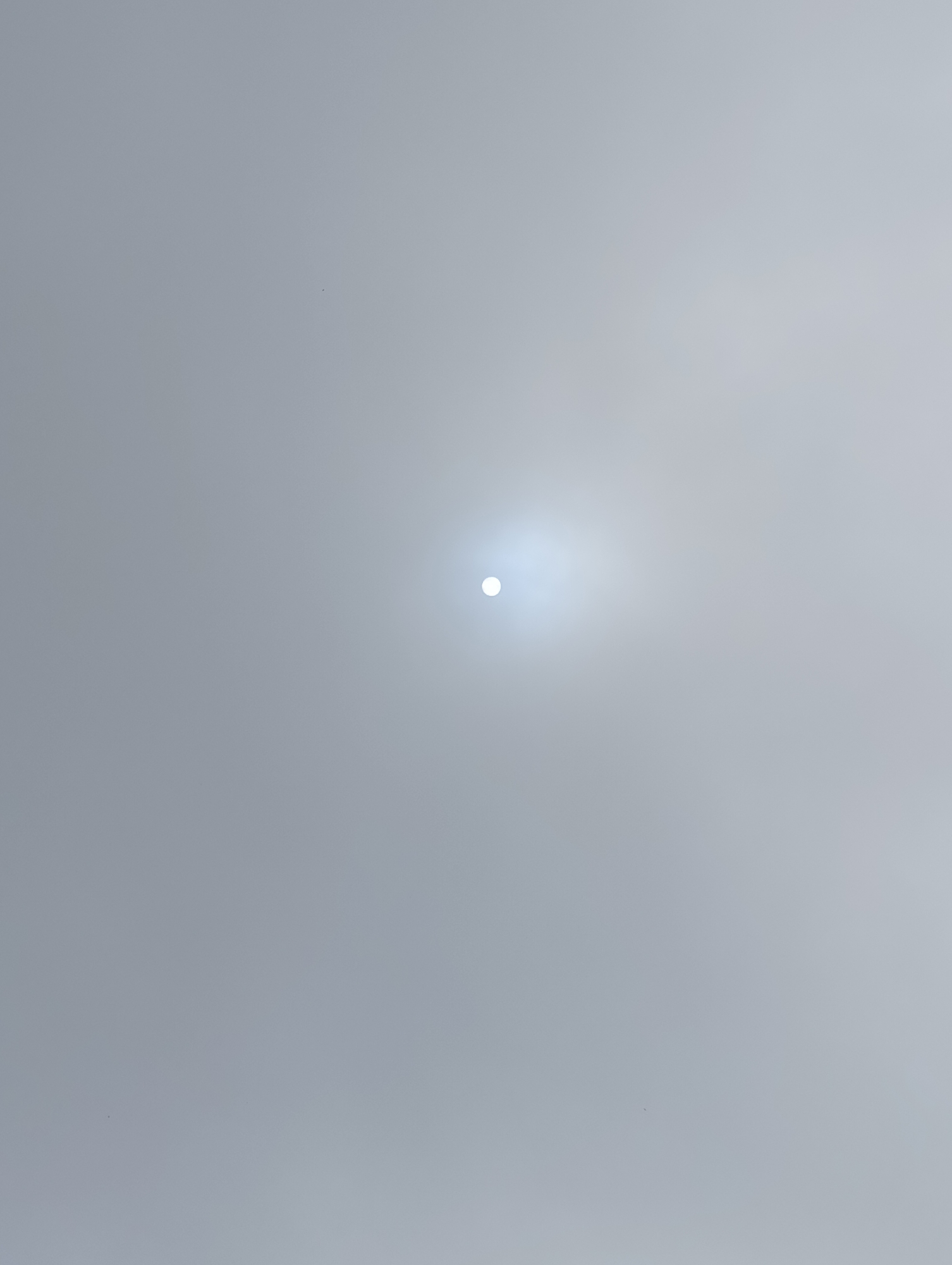 The image displays a bright, luminous orb, likely the sun, positioned centrally against a hazy, overcast sky. The sky is a uniform pale gray, suggesting diffuse light and possibly fog or a heavy layer of clouds. There are no discernible landmarks or objects in the sky or on the ground. The overall impression is one of a bright but obscured celestial body. There is no information in the image to suggest a specific location in Siumsa, India.