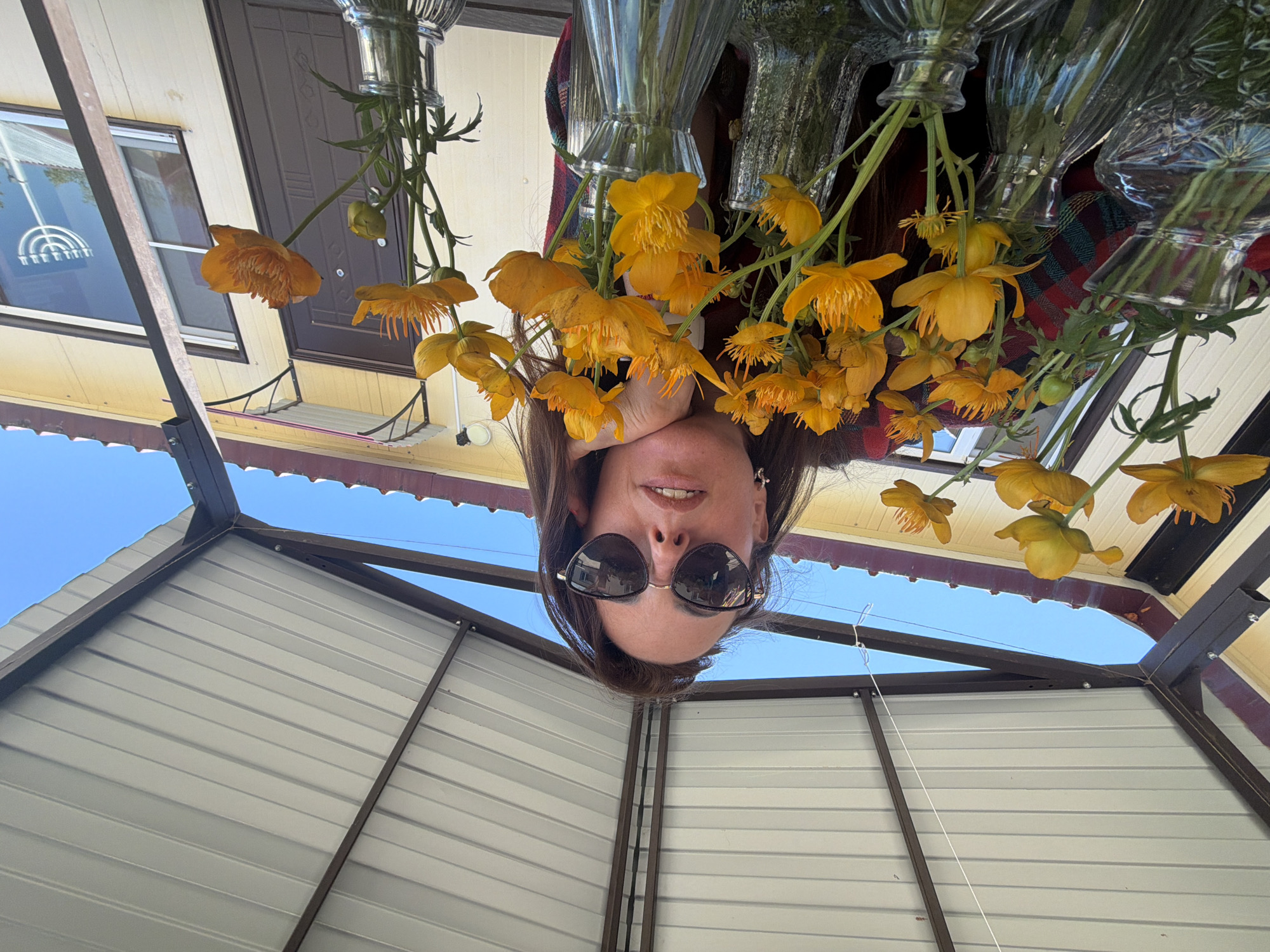 A woman wearing sunglasses and a red and black plaid shirt is leaning on her hand, posing with a bouquet of yellow flowers in glass vases. She is under a white corrugated metal awning supported by brown metal beams. Behind her, a yellow building with brown trim and a brown door is visible. A window on the building reflects a menorah. The sky is clear and blue. The overall scene suggests an outdoor setting, possibly a yard or patio. The location is identified as Birobidzhan, Russia, though this detail is not visually verifiable from the image itself.