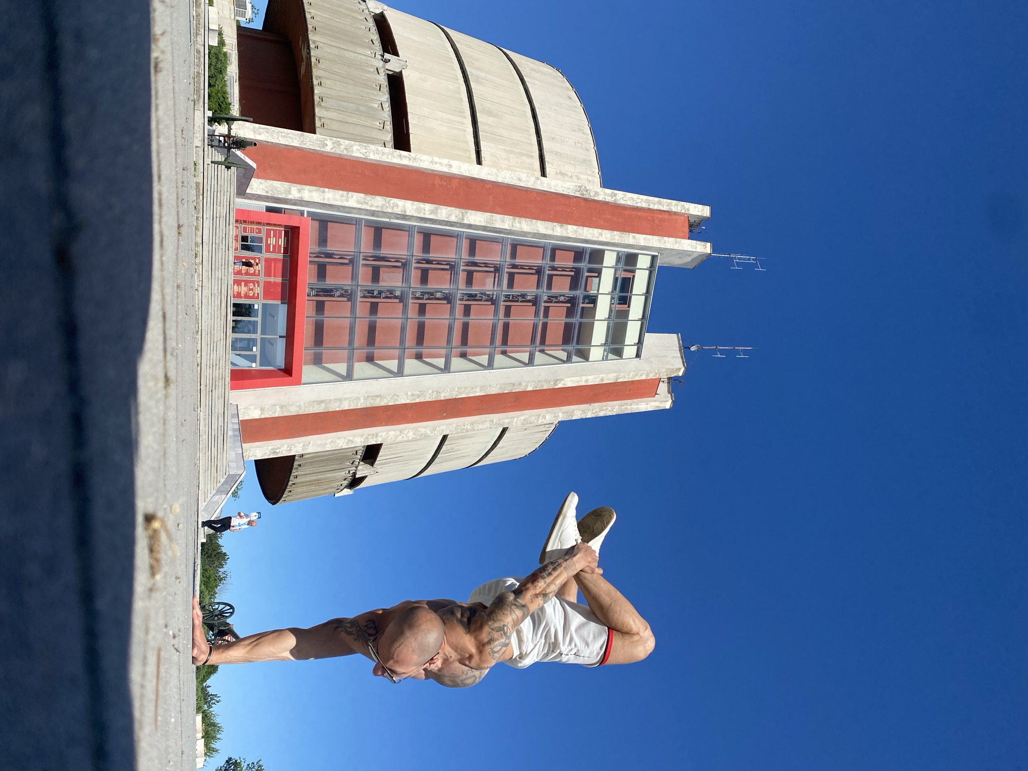 A bald man with tattoos is performing a handstand on a concrete ledge. He is wearing white shorts and sneakers. In the background is the Pleven Panorama building in Pleven, Bulgaria, with its distinctive concrete structure and red and white facade. The sky is clear and blue. Another person and a cannon are visible in the background, near the base of the building.