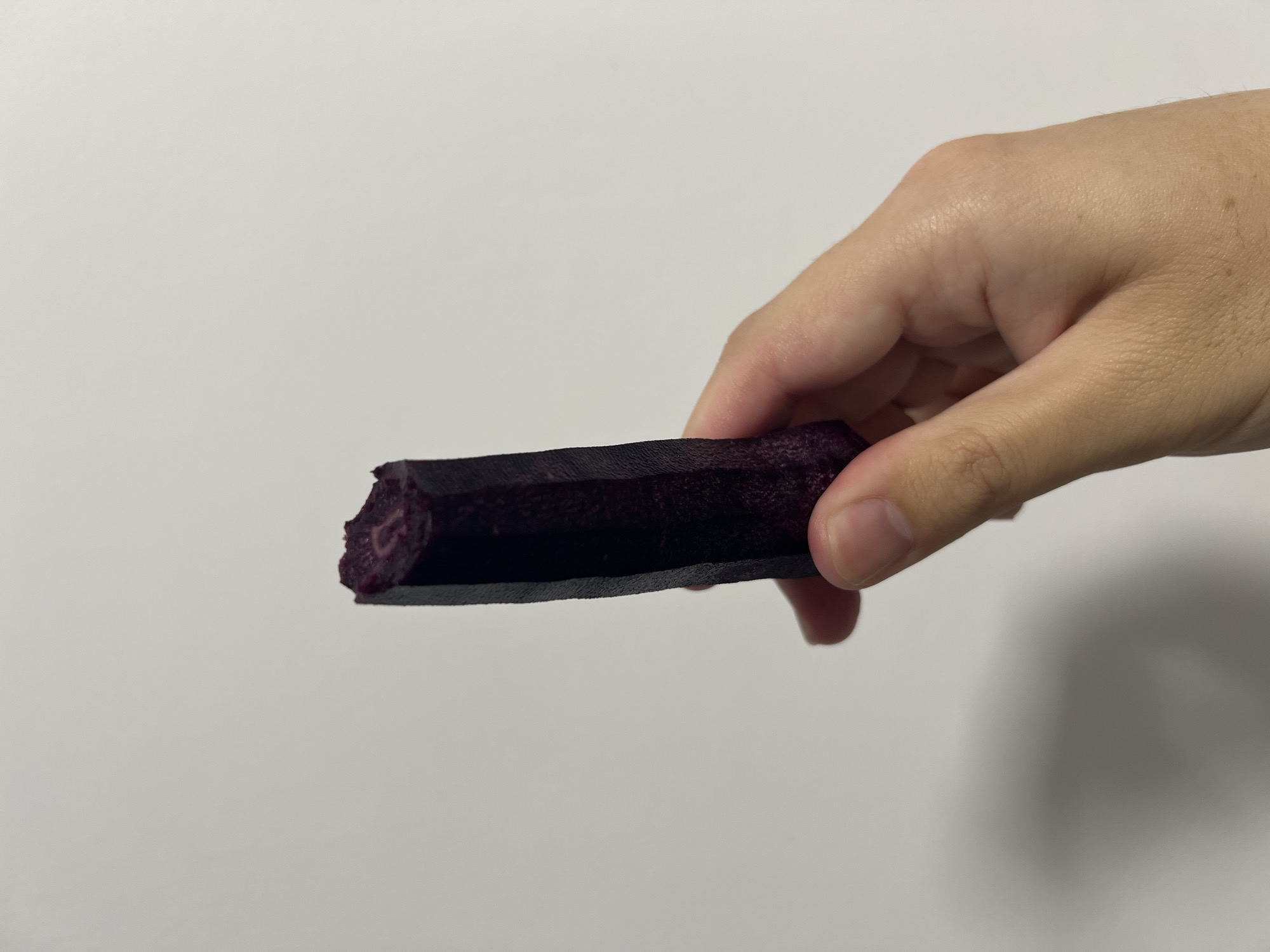 The image displays a close-up of a person's hand holding a dark purple, cylindrical object against a plain, light grey background. The object appears to be an edible item, possibly a piece of fruit, vegetable, or a snack bar, and has an irregular, somewhat textured surface. One end of the object shows a bite or break, revealing a reddish-purple interior with a fibrous or pulpy texture. The person's fingers are wrapped around the object, holding it horizontally across the frame. The lighting is bright and uniform, suggesting an indoor setting. No specific location cues, time of day, weather, or discernible emotions are evident from the image itself, apart from the provided context that it is from Mogán, Spain. There is no visible text.
