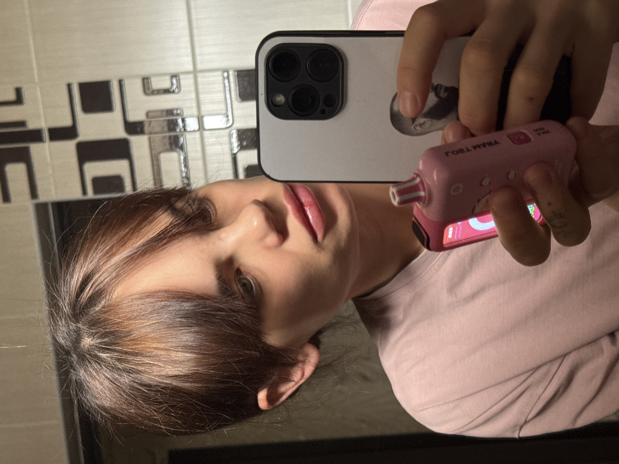 The image shows a person taking a selfie in what appears to be a bathroom. The person is holding a smartphone and a pink handheld device. The background features a tiled wall with a patterned design. The person's hair is brown with highlights, and they are wearing a light pink shirt. There is no information present in the image to suggest the city or country where it was taken.