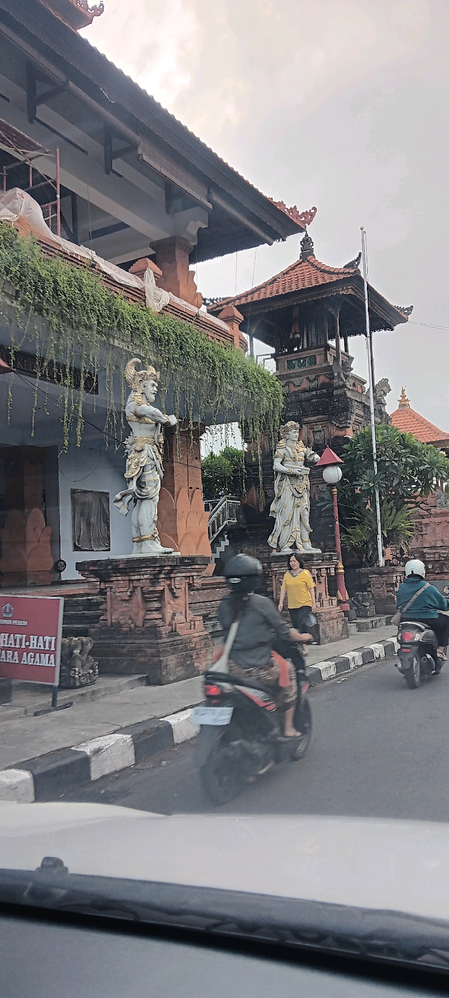 In Denpasar, Indonesia, a scene unfolds with traditional Balinese architecture as a backdrop. Two large, ornate statues of guardian figures stand guard on a raised brick platform in front of a building with a dark-tiled roof and lush green vines cascading from its overhang. The building itself features a white facade and intricate brickwork at its base.
To the right of the statues, a multi-tiered structure with a red-tiled roof, characteristic of Balinese temples, rises. A lamppost with a white globe stands between the statues and this structure.
The street in front of the building is paved, with a distinct black and white striped curb. Several people are present. On the street, a woman wearing a yellow shirt is walking. Two individuals on motorcycles are also present; one, wearing a gray shirt and black helmet, is in the foreground, while another, wearing a teal jacket and white helmet, is further down the street.
A red sign with white text, possibly a warning or informational notice, is visible on the left side of the image, placed near the base of the statues. The sky above is overcast. The perspective suggests the image was taken from within a vehicle, likely looking out through the windshield.