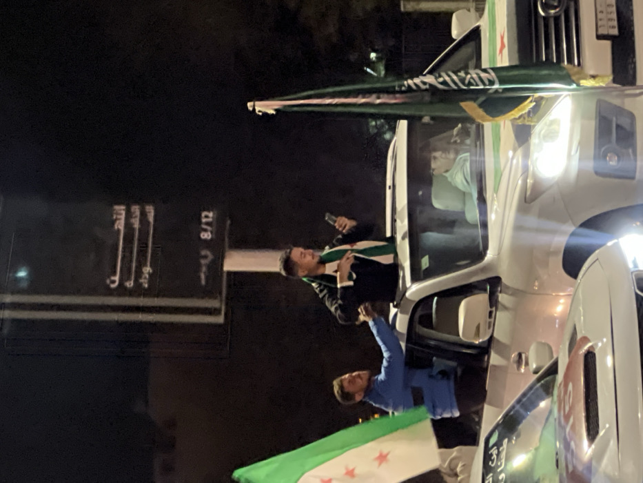 The image captures a nighttime scene, likely on a street or road in Al-Qanawat Municipality, Syria. Several men are engaged in what appears to be a celebratory or demonstrative event, leaning out of the windows of white vehicles, which resemble SUVs or vans.
In the center, a man with a beard, wearing a black jacket and a scarf patterned with the Syrian opposition flag (green, white, black stripes with three red stars), leans out of a vehicle, holding a mobile phone. Below him, another man in a blue shirt also leans out, holding a larger Syrian opposition flag that extends downwards. On the front of the vehicle they are leaning from, a prominent green flag with white Arabic calligraphy, likely depicting the Shahada ("La ilaha illallah, Muhammadun rasulullah"), is mounted. A small emblem resembling the Syrian opposition flag is also visible on the side of this vehicle near the headlight. Another white vehicle is partially visible in the foreground, showing a red square with the number "10" on its side.
The time of day is clearly night, as indicated by the dark background and the illuminated headlights of the vehicles. In the background, a dark street sign is visible with Arabic text. Parts of the text readable include "الله" (Allah), "أكبر" (Akbar), "وداد" (Widad), and the numbers "8/12." The overall atmosphere appears enthusiastic and active.