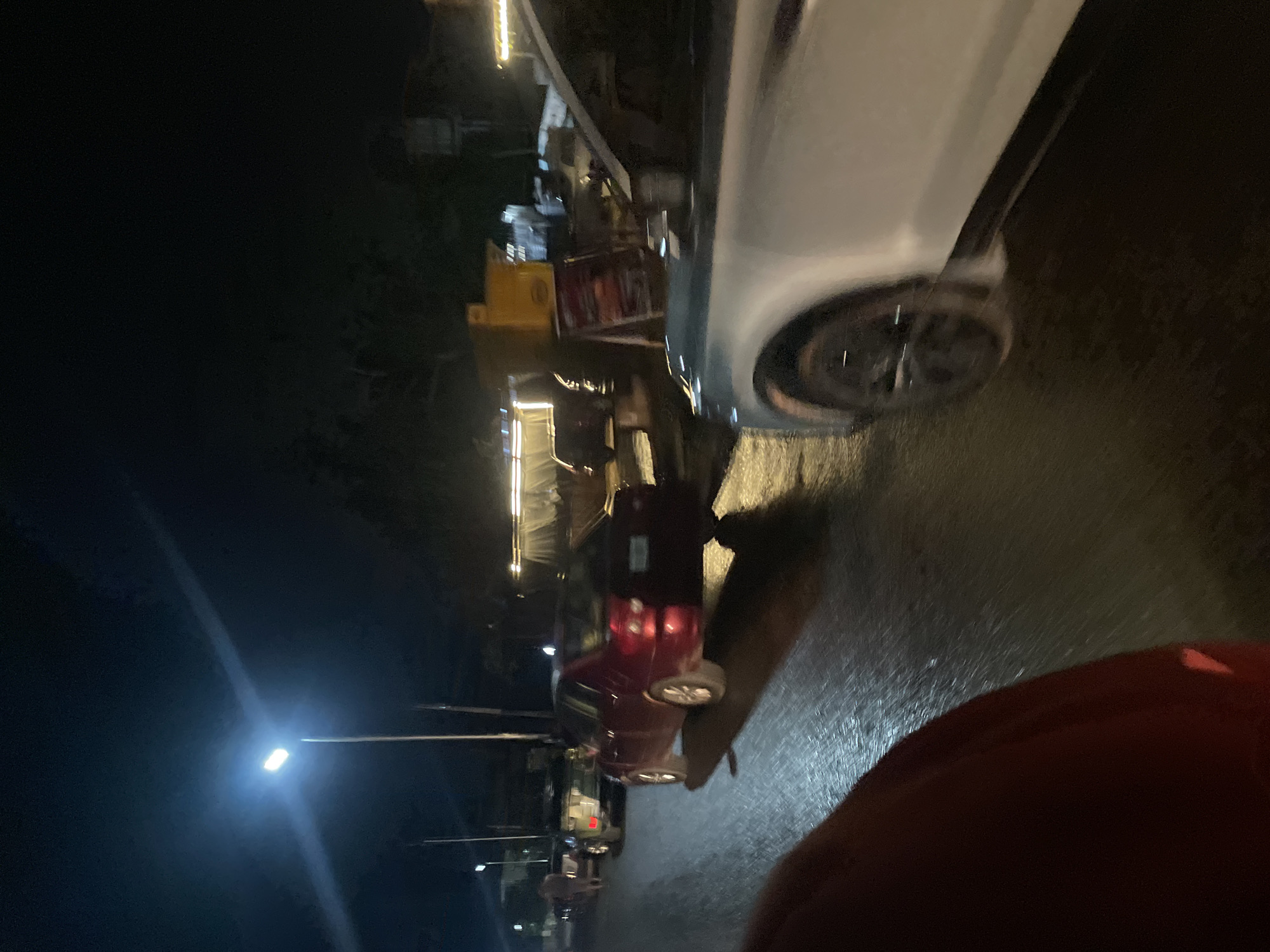 The image captures a nighttime street scene in Abuja, Nigeria, featuring a lively urban environment illuminated by artificial lights. The perspective is from a low angle, with a person's shoulder, wearing a red garment, partially visible in the bottom right foreground.
The scene is dominated by several parked cars along a wet or reflective road surface, suggesting recent rain or reflective tarmac. On the right, a silver or light grey car's front side and wheel are prominently featured. Further into the mid-ground, a red car is clearly visible, parked with its rear facing the viewer. Other vehicles are faintly discernible further down the street.
The left side of the street is lined with brightly lit structures, likely market stalls or small commercial establishments, which contribute to the overall illumination of the area. A prominent street light glows on the far left, casting light onto the dark surroundings. The overall impression is of a bustling commercial street at night. No identifiable text is visible within the frame.