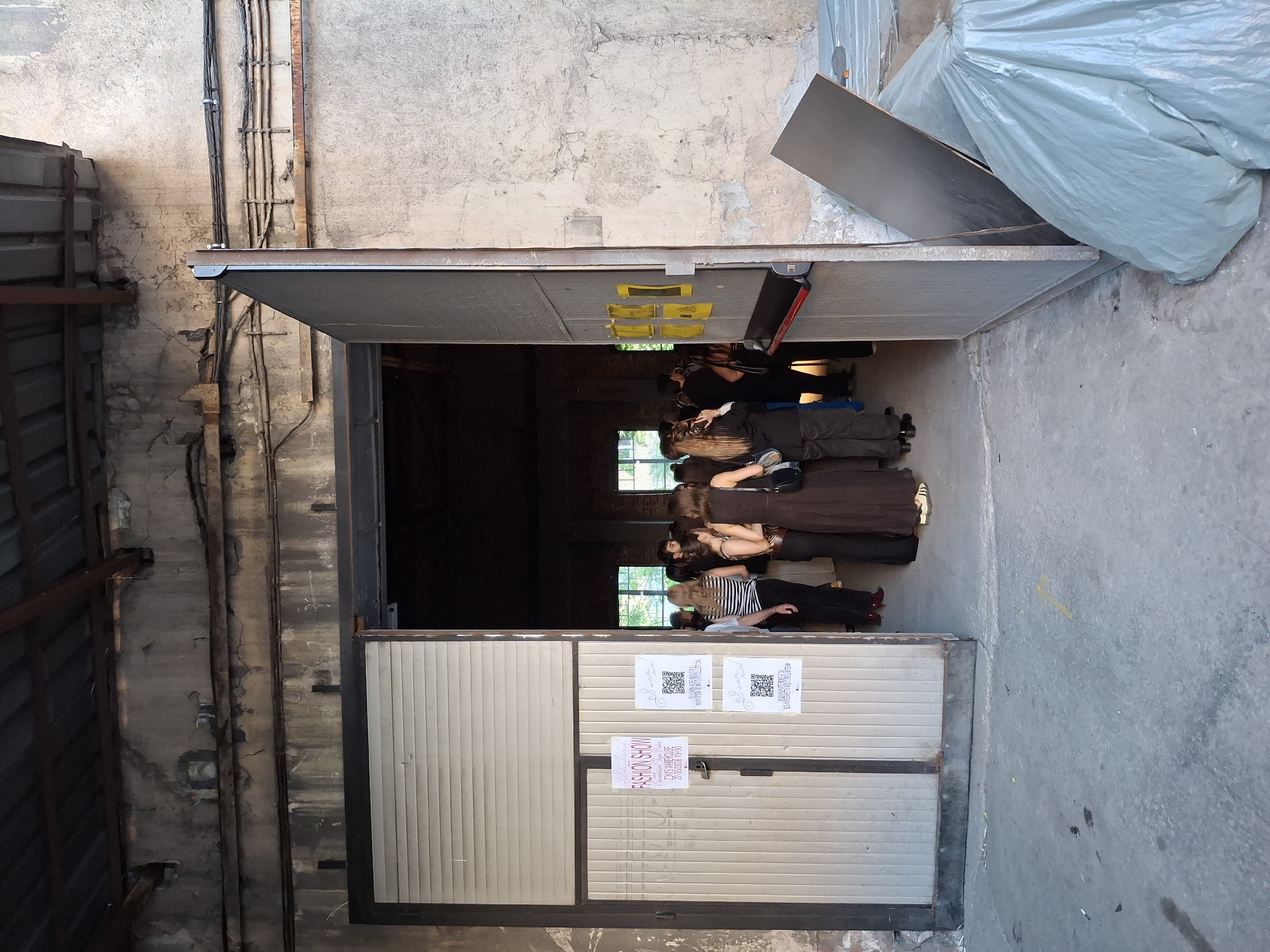 The image shows a scene inside a building, seemingly a warehouse or industrial space, in Unknown, Unknown. A group of people are standing in line, presumably waiting to enter or view something. They are mostly dressed in casual clothing.
The building has a large, partially open metal door, which is the main focal point of the image. The door has posters or signs attached to it, including one with a "FASHION SHOW" advertisement and QR codes. The building's interior is dimly lit, but two windows in the back are visible, allowing some natural light to enter.
The walls are made of concrete and appear aged or weathered. Various pipes and conduits are visible running along the walls. Outside the building, there's a pile covered with a light blue tarp. The overall environment seems to be an urban or industrial setting.