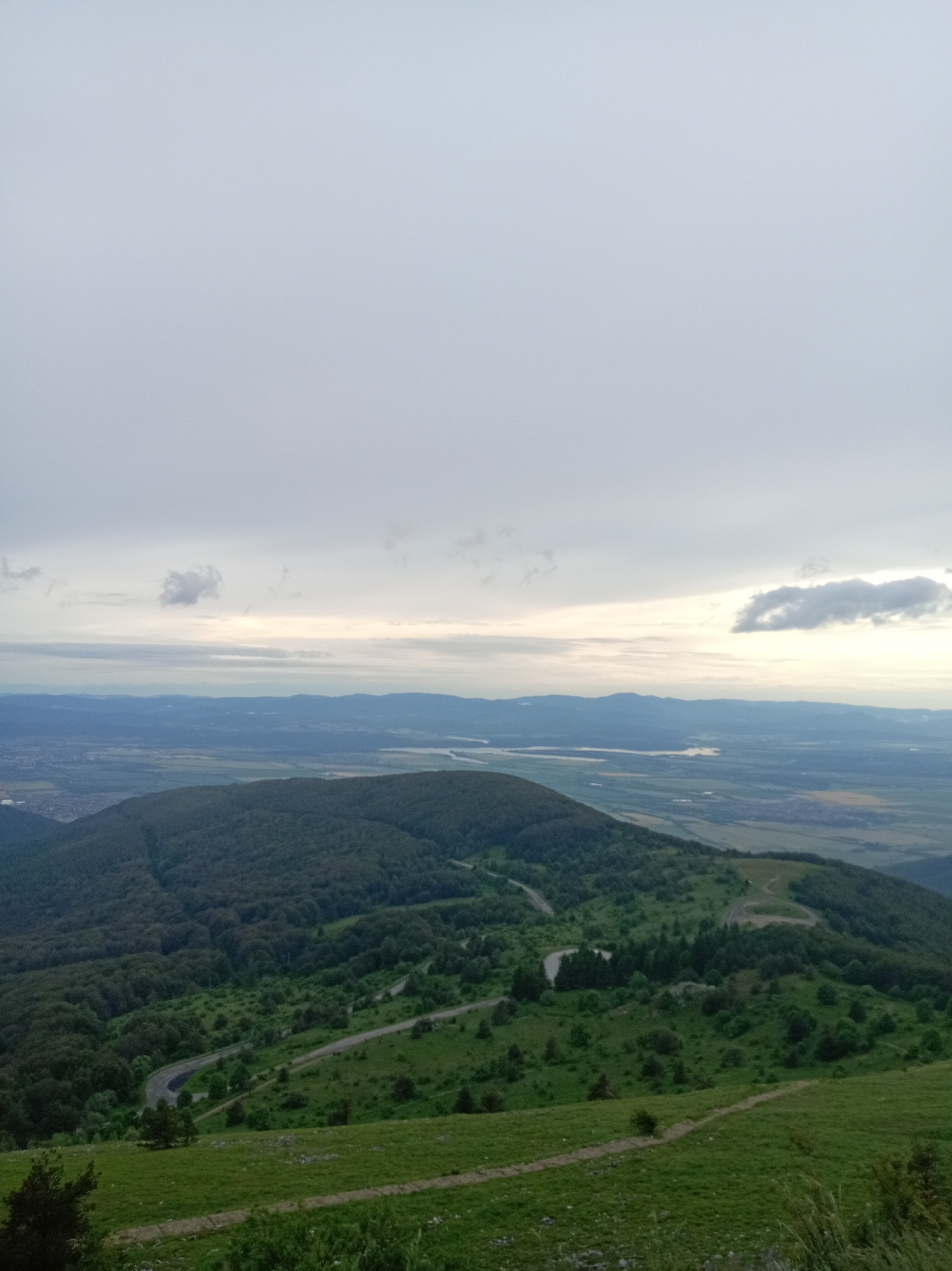 This is an eye-level, wide-angle, outdoor shot taken from a high vantage point, looking down and out over a vast landscape. The sky occupies the top half of the frame and is filled with a thick, overcast layer of pale gray clouds, with hints of lighter tones suggesting a muted sunrise or sunset. A few wispy, darker clouds are scattered across the sky.
Below the sky, a hazy, multi-layered vista unfolds. The immediate foreground, at the bottom of the image, is a lush green grassy slope. A narrow, light-colored path or trail winds across this slope from the right side of the frame.
Further into the mid-ground, a dense, dark green forest covers a rolling hillside. A winding road, appearing as a thin gray line, carves its way through this wooded area. The forest extends and then transitions into a more varied landscape.
The background reveals a broad valley with a patchwork of fields and smaller wooded areas. In the far distance, a body of water, possibly a river or lake, is visible, reflecting the muted light of the sky. A hazy outline of distant mountains or hills forms the horizon. The overall impression is one of a natural, expansive, and somewhat subdued environment under a cloudy sky. There are no people or animals visible in the image.