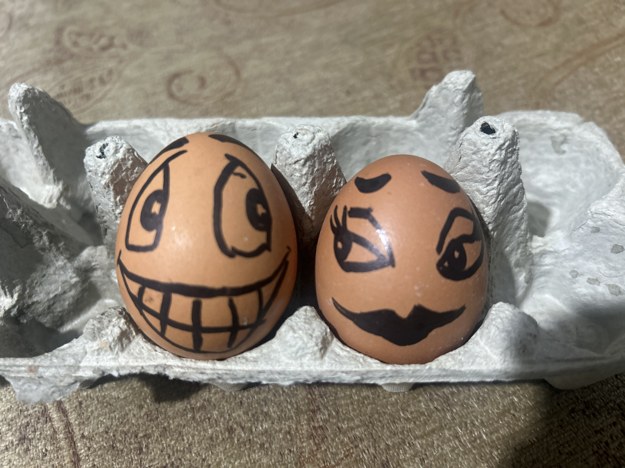Two brown chicken eggs are in a grey cardboard egg carton. Each egg has a face drawn on it with a black marker. The egg on the left has wide, cartoonish eyes and a large, toothy grin. The egg on the right has one eye winking and large, smiling lips. The egg carton is on a surface with a textured, beige and brown pattern. The location is Unknown, Unknown.