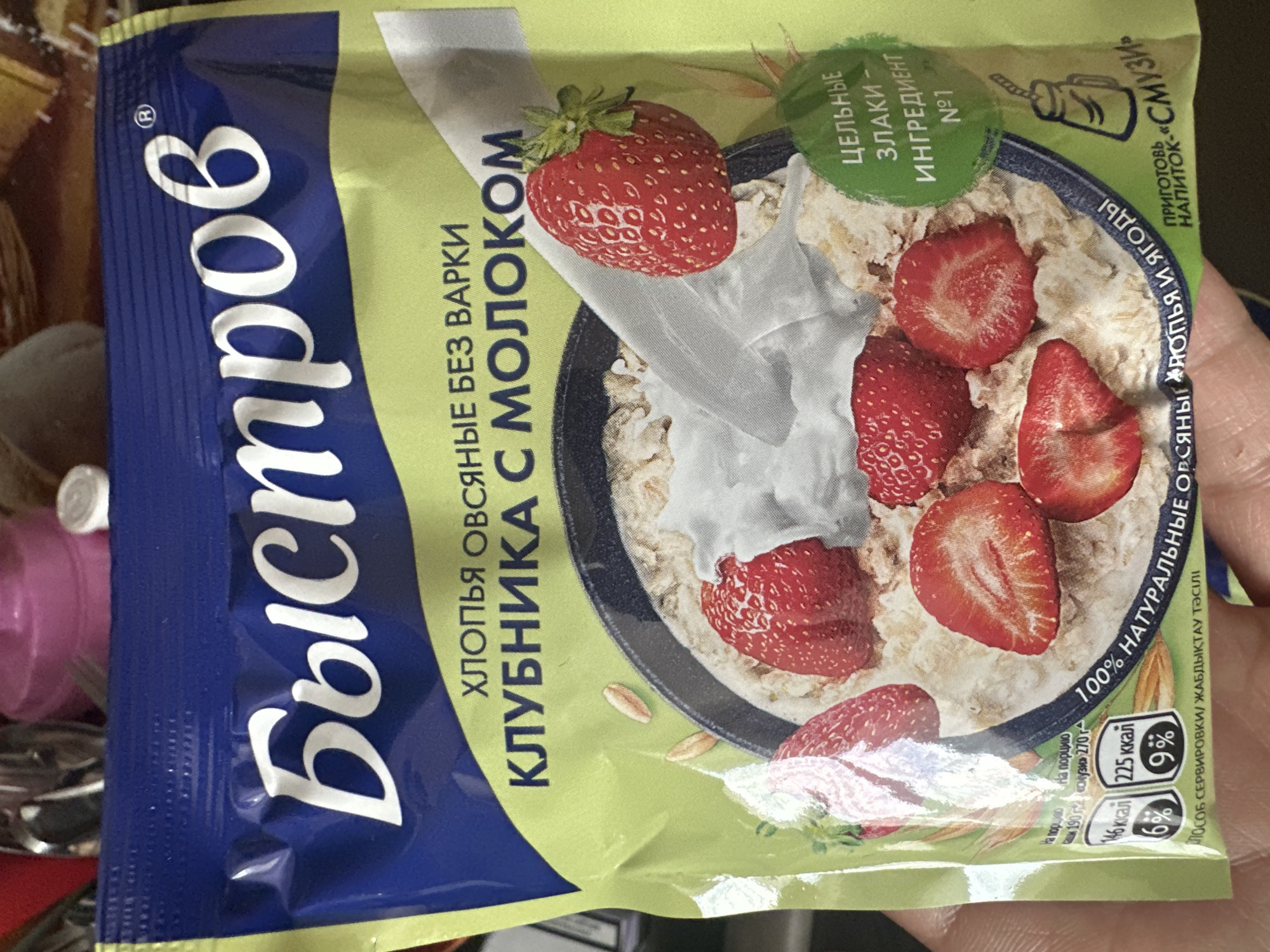 The image shows a close-up of a package of "Быстров" brand instant oatmeal with strawberries and milk. The packaging is primarily blue and lime green, featuring an illustration of oatmeal in a bowl with strawberries and cream. Text on the package is in Russian and indicates "Oatmeal without cooking," "Strawberry with milk," and "100% natural oatmeal with strawberries and berries." There is also a circular green graphic that reads "Whole grains - Ingredient No. 1." Nutritional information and serving suggestions are also visible. A person's hand is holding the bottom of the package, obscuring a portion of the lower right corner. The background is blurred and appears to be an indoor setting with indistinct objects, possibly kitchenware. There is no visible information that would place this image in Novoaltaysk, Russia.