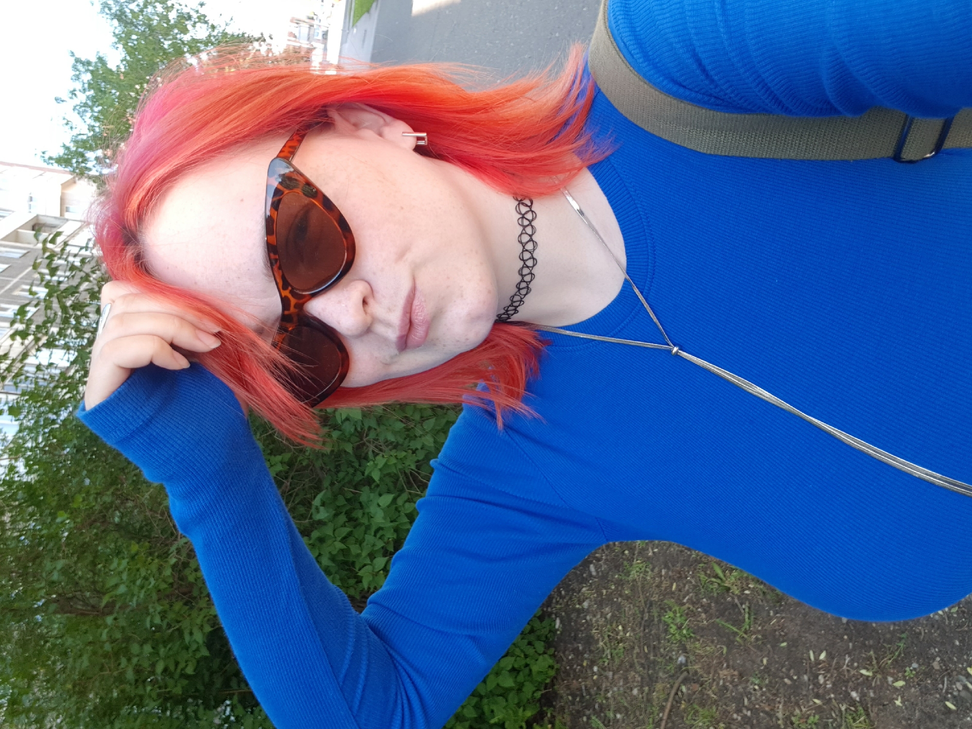 A woman with dyed red hair and freckles poses for a photo outdoors in the city of Unknown, Unknown. She is wearing brown sunglasses and a long-sleeved blue shirt, with a thin silver necklace and a black choker. Her arm is raised, with her hand near her forehead. A green backpack strap is visible over her shoulder. The background includes green bushes and a building. The ground is a mix of grass and dirt.