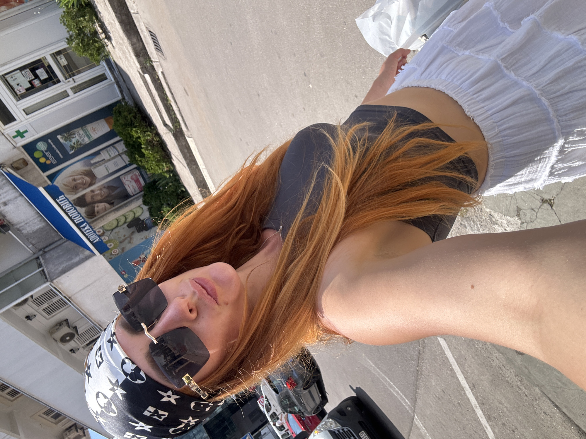 This media file is a self-portrait taken outdoors, likely in a sunny urban environment. The main subject is a person with long, reddish-blonde hair, wearing dark sunglasses with square frames, a dark tank top, a white tiered skirt, and a black and white patterned headscarf. Their arm is visible, and a small mole can be seen on their upper arm. The person is holding a white plastic bag in their right hand.
The background shows a street scene with parked cars and a building that appears to be a pharmacy or medical facility, indicated by a green cross logo and signage with Cyrillic text. Some of the visible text on the signage appears to read "АПТЕКА" (Apteka, meaning pharmacy) and includes images of cosmetic products. The overall setting suggests a daytime activity, possibly shopping or walking in a town. The lighting indicates it is daytime, and the bright sunlight suggests fair weather. The location context of Budva, Montenegro, aligns with the visual cues of a European urban environment with such a building and street.