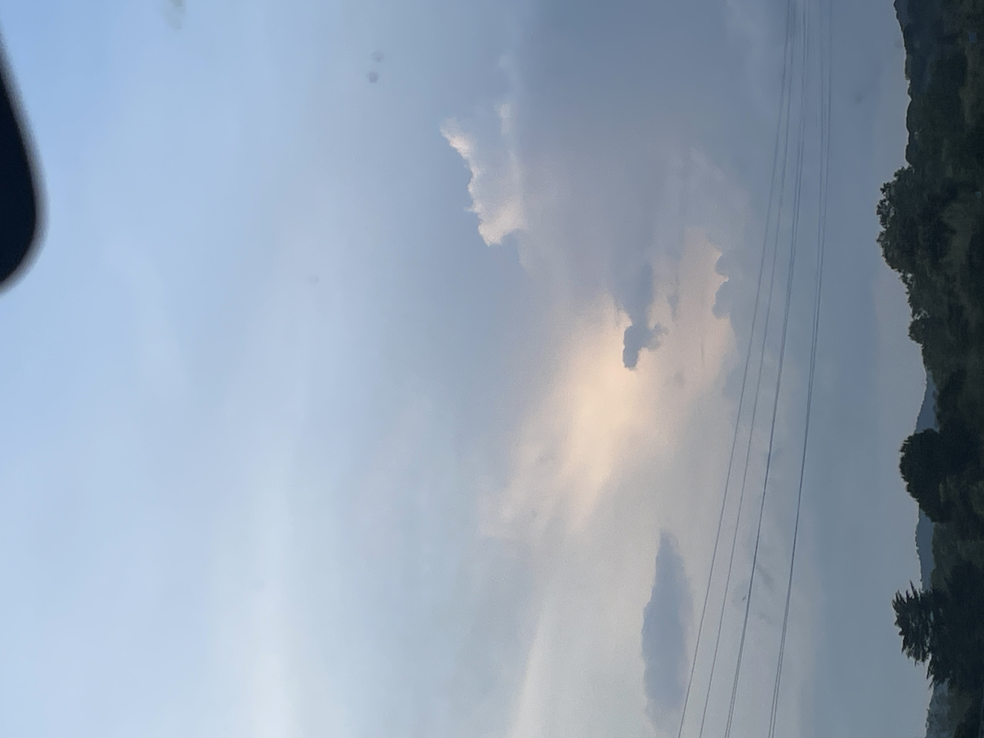 This image, when viewed in a horizontal orientation (rotated 90 degrees counter-clockwise from its current state), depicts a vast sky over a natural landscape in Argentona, Spain. The sky dominates the frame, transitioning from a clear, light blue on the left to a hazier, cloudier area towards the right. Several cloud formations are visible, with one prominent, illuminated patch near the center exhibiting a soft, yellowish-orange glow, indicative of either late afternoon sunlight breaking through or an early morning sunrise.
Numerous thin, dark lines, consistent with power lines or utility wires, stretch horizontally across the middle and upper-right sections of the sky. Along the bottom edge of the image, a dark, undulating silhouette of dense trees and vegetation suggests a forested hillside or mountainous terrain. In the top-left corner, a blurred, dark, curved object is visible, likely a part of a car's interior such as a dashboard or rearview mirror, implying the photograph was taken from within a vehicle.
The overall atmosphere suggests an overcast or partly cloudy day with soft, diffused light. There are no people or specific events occurring. No visible text is present in the image.