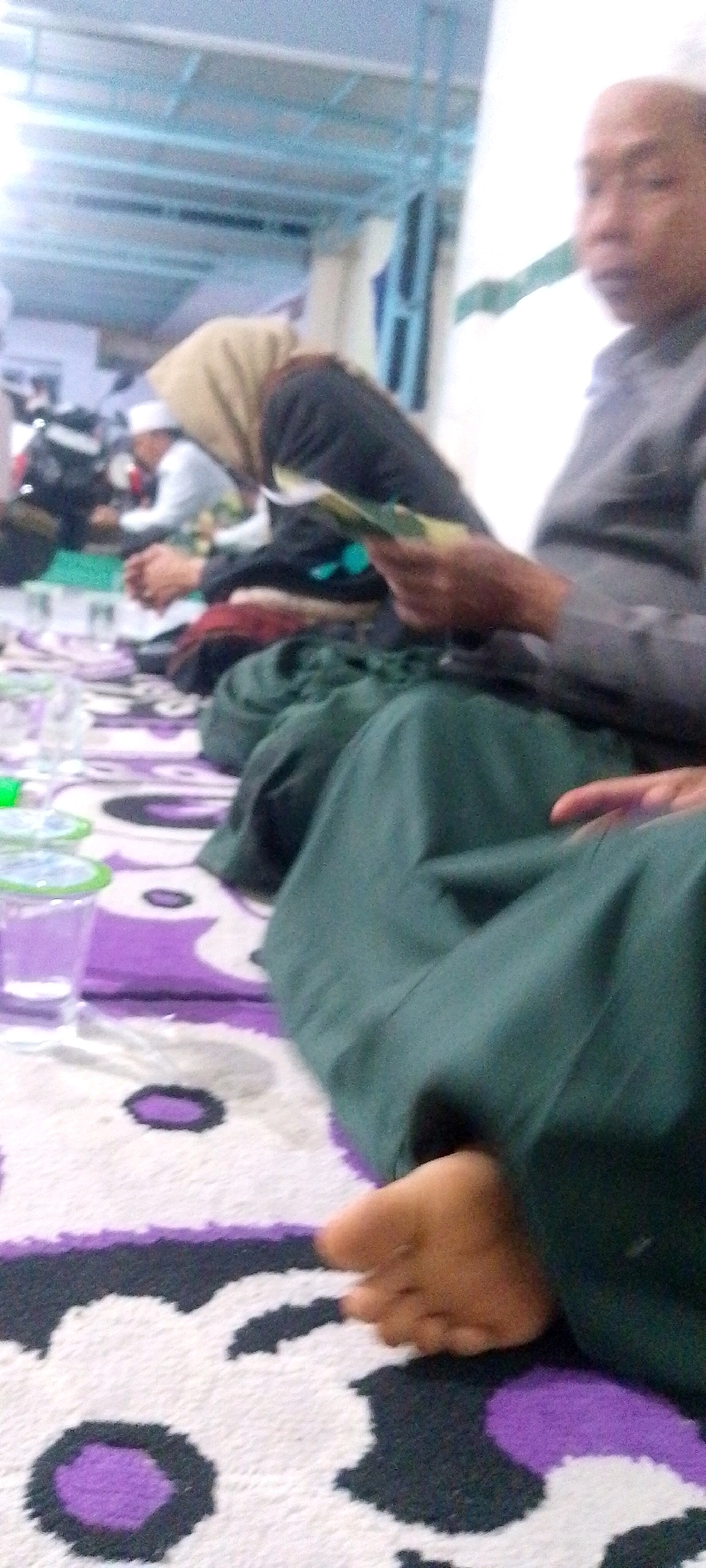 A medium-shot, indoor, slightly blurred, low-angle photograph captures a group of people sitting on the floor. The floor is covered with a white, purple, and black patterned rug. In the foreground, a person's bare feet are visible, resting on the rug. A dark green garment is draped over the legs of this person. To the left, several clear plastic cups with green lids are placed on the rug, one filled with a light-colored liquid. In the background, other individuals are seated, their faces largely out of focus. One person in the middle ground appears to be wearing a white cap and a light-colored shirt. Another person is wearing a dark head covering and a black garment. The environment suggests an informal gathering or religious event, possibly a mosque or community center, with a ceiling structure visible in the upper portion of the frame. The lighting is bright and appears to be from overhead.