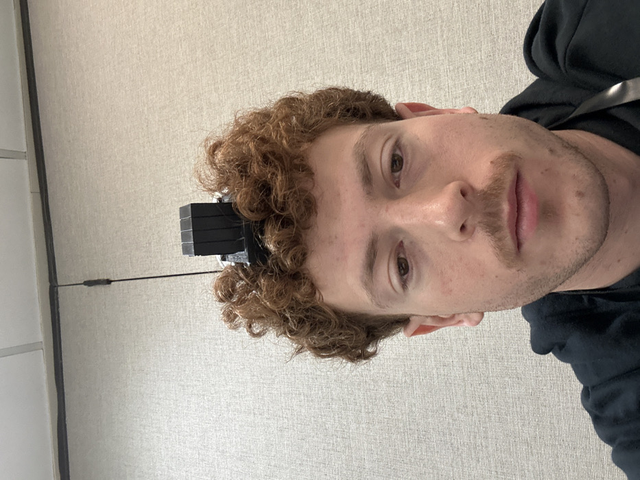 An image depicts a close-up, head-and-shoulders view of a young adult male, appearing Caucasian, positioned indoors. The subject has reddish-brown, curly hair and a mustache. His eyes are dark and gaze directly forward. A black tefillin shel rosh, a square box with a multi-segmented rectangular projection, is worn on the top of his head, partially obscured by his hair. He is wearing a dark-colored hooded garment, and a dark lanyard is visible over his right shoulder. The background consists of a light-colored, textured wall. Above the wall, a section of a suspended acoustic ceiling with white tiles and dark grid lines is visible. A thin, dark cable or cord is present vertically against the upper portion of the wall. No specific location within Fairfield, United States of America, is identifiable from the visible details.