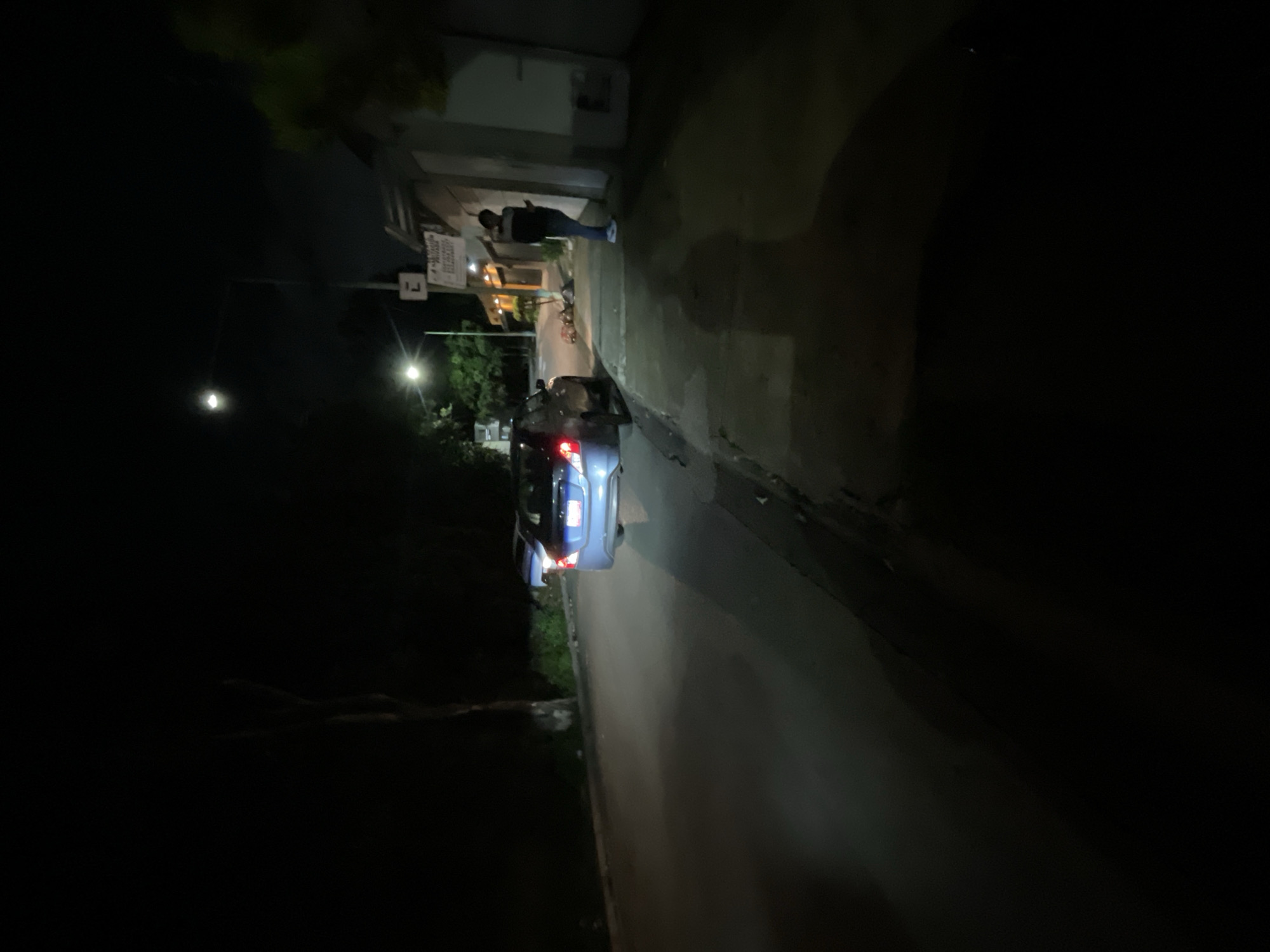 A blue sedan is parked on the side of a dark, paved road at night. The car's taillights are illuminated. A person, wearing a dark jacket and jeans, stands on the sidewalk next to a building. The building appears to be a storefront with a sign. Streetlights illuminate parts of the scene, casting long shadows. The environment suggests a residential or commercial street in Huixquilucan de Degollado, Mexico, given the presence of a building with a Spanish sign and a common vehicle model. The overall lighting conditions indicate nighttime.