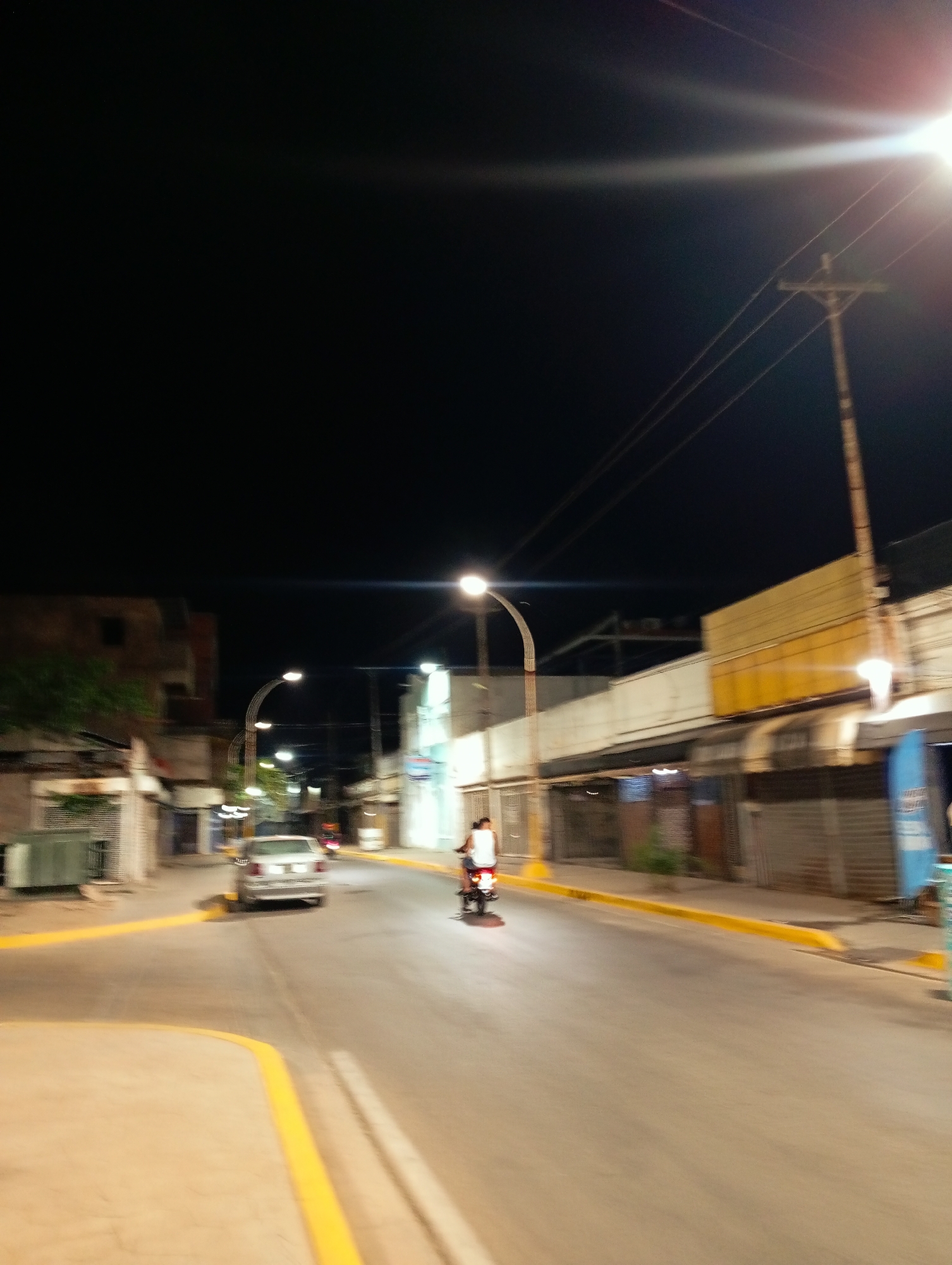 A nighttime street scene in Juan Griego, Venezuela shows a paved road illuminated by streetlights. A silver sedan travels in the same direction as a person on a motorbike. Buildings line both sides of the street, with storefronts closed by metal roll-up doors. A yellow curb is visible along the right side of the street. A wooden telephone pole and wires are visible on the right side of the street.