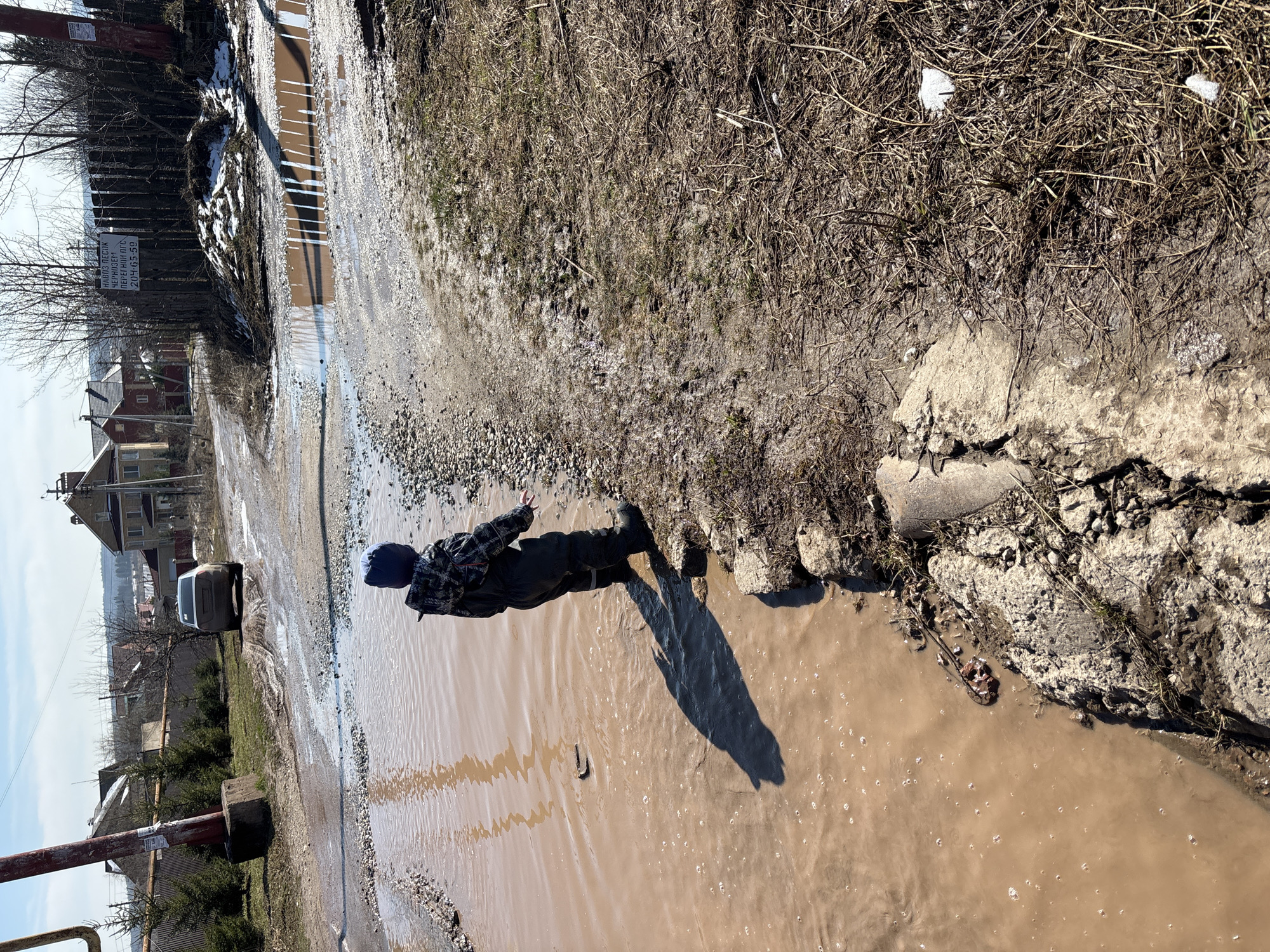 A person stands in a large puddle of muddy water on a street in Ферма, Russia. The water covers most of the road, reflecting the sunlight. To the left, a car is partially visible driving through another section of the flooded street. Buildings and trees line the road in the background. The person is wearing dark clothing and a hat and appears to be gesturing with one hand. The edges of the flooded area are bordered by dirt, gravel, and some vegetation.