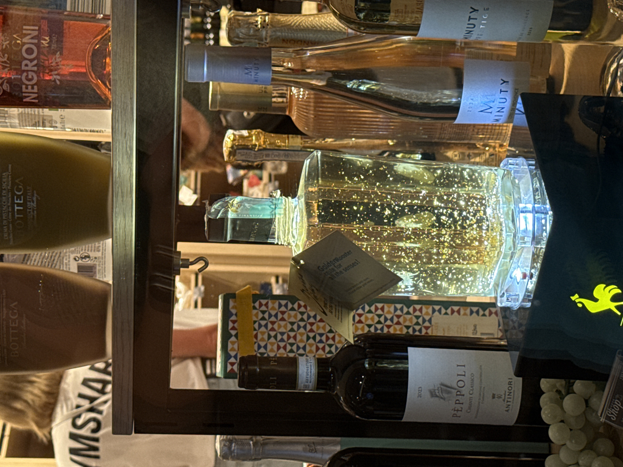 The image shows a display of bottles in what appears to be a liquor store or a duty-free shop. The bottles are arranged on shelves and a counter. In the foreground, there is a bottle with a clear liquid containing gold flakes, illuminated from below by blue lights. To its left is a bottle of red wine labeled "PEPPOLI CHIANTI CLASSICO" from Antinori, with the year 2023 visible. Behind the gold-flecked bottle are several other bottles, including what looks like a bottle of rosé wine with a label that includes "MINUTY" and "COTES DE PROVENCE," dated 2024, and another wine bottle also with a "MINUTY" label. Further back, there are more bottles, some with metallic foil wrapping. In the background, a person with blonde hair wearing a white t-shirt with the word "YMSHAP" in black letters is visible, as are shelves with more bottles and containers. A part of a sign with the word "BOTTEGA" and "NEGRONI" is visible on the top shelf. There are also some grapes in the lower left corner of the image. The overall lighting is dim, with spotlights illuminating the products. The setting is likely commercial. Without further information, pinpointing the exact location in Budva, Montenegro is not possible.