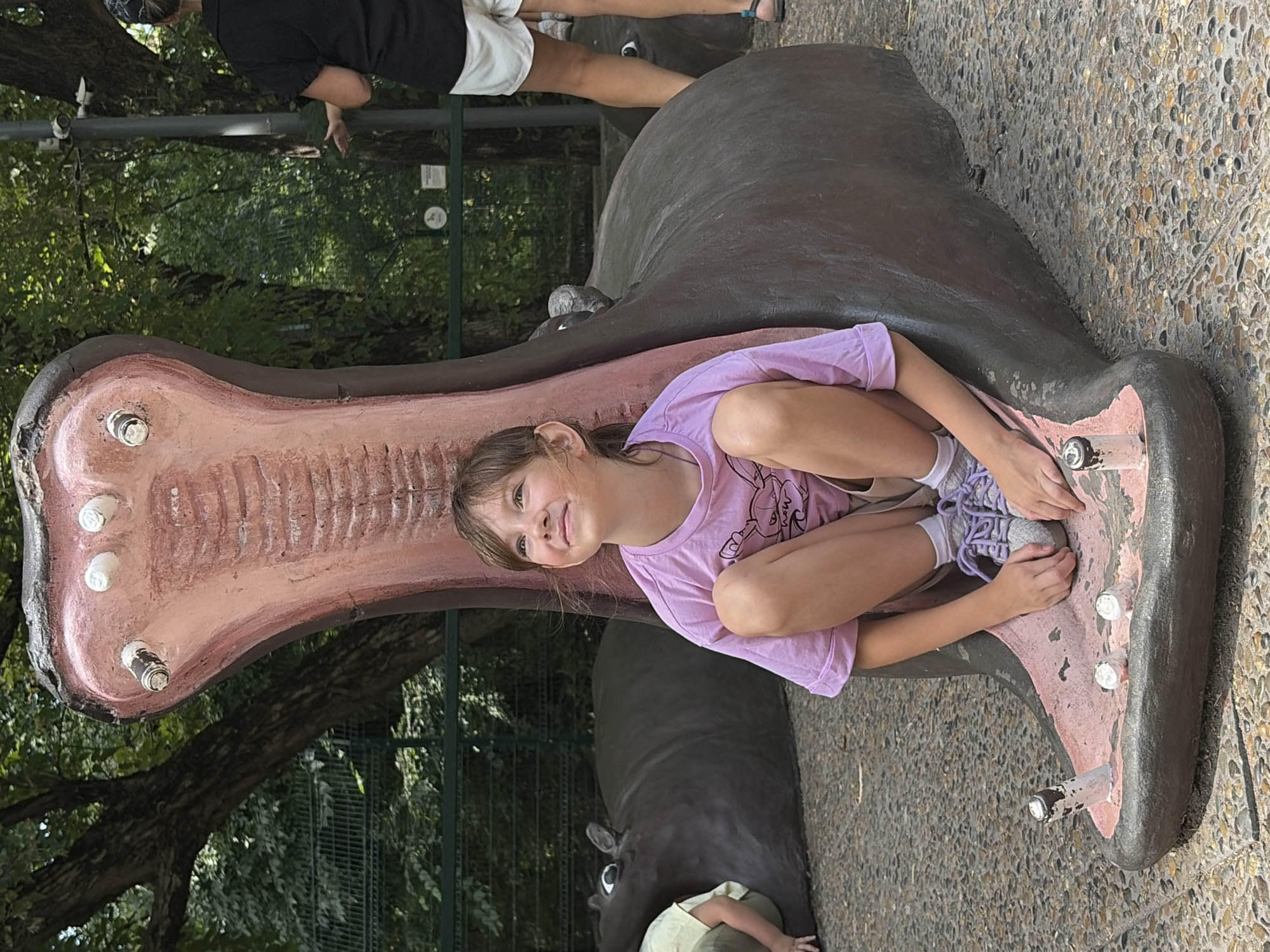 A young girl with light brown hair is sitting inside the open mouth of a large, stylized hippopotamus sculpture in an outdoor setting. The sculpture is dark gray with a pink interior, featuring large, white, bulbous teeth. The girl is wearing a light purple t-shirt with a graphic print and beige shorts, with white socks and purple sneakers. She is looking upwards and to the right with a smile. In the background, another hippopotamus sculpture is visible to the left, partially obscured, with a child in a light green shirt visible sitting on it. Further in the background, trees and foliage are present, along with a metal fence and a lamppost with security cameras. A person wearing a black top and white shorts is standing to the right, facing away from the camera. The ground is covered with gravel. This scene is located at the Rostov Zoo in Rostov-on-Don, Russia.