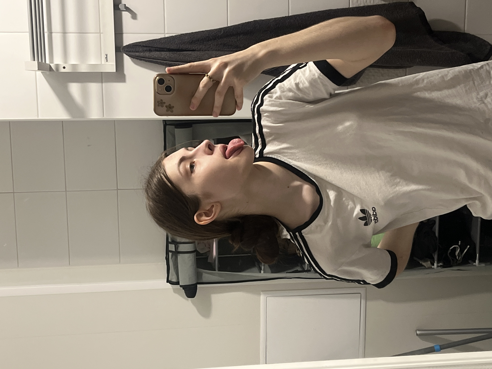 The image is a mirror selfie taken indoors, likely in a bathroom setting in Saint Petersburg, Russia. The entire scene is rotated 90 degrees counter-clockwise.
The primary subject is a young person, appearing to be female, with light skin and dark brown hair styled in a bun or ponytail at the back of her head. She is wearing a white t-shirt with black trim on the collar and sleeves, accented by three black stripes running down the shoulders. A black Adidas trefoil logo is visible on the right side of the chest, appearing as "bibo" due to the image's rotation. The person holds a light brown or gold-colored smartphone in her left hand, which has a case featuring two light-colored floral or leaf designs on the back. Three camera lenses are visible on the phone. A beaded ring is worn on one of her fingers. Her head is tilted back, and her tongue is playfully sticking out, conveying a casual and expressive mood.
The background features white rectangular wall tiles. A white, slatted object, possibly a shelf or radiator, is mounted on the tiled wall. Faint text "Radia..." can be seen on this object. A dark grey or black towel is draped over a fixture in the upper right portion of the background. Below the person's head, a black mesh hanging organizer contains several indistinct items. A white panel, possibly a door or cabinet, is visible in the lower part of the reflection.