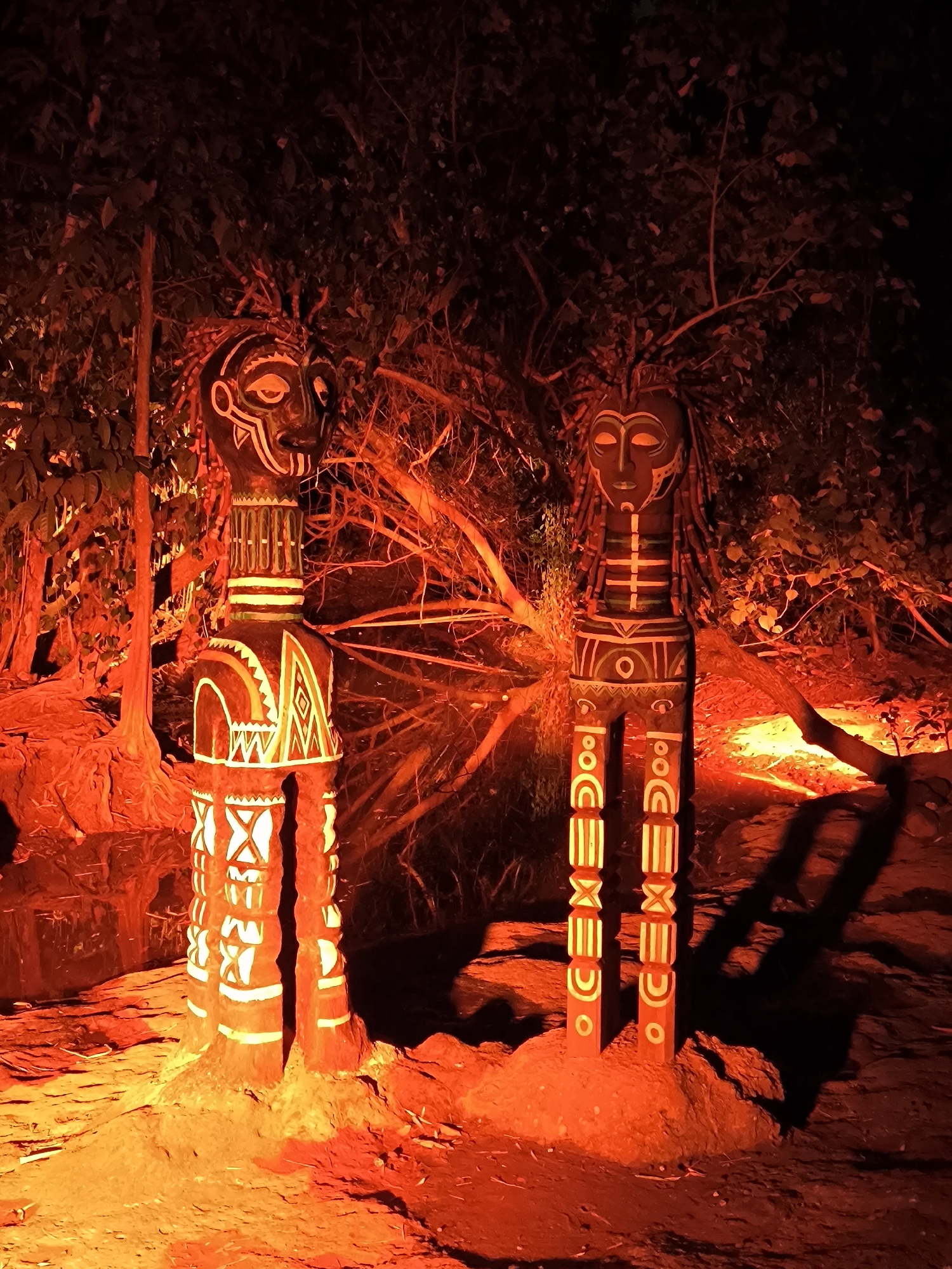 The image depicts two tribal statues illuminated by warm, orange lighting, set against a backdrop of dark foliage and trees. The statues, one on the left and one on the right, are carved from wood and adorned with intricate white patterns. The statue on the left has a broader torso and legs, while the statue on the right is more slender. Both statues have distinct facial features and headdresses. The ground around the statues appears to be rocky and uneven, with shadows cast by the statues and surrounding vegetation. The overall atmosphere is mystical and evocative, suggesting a ritualistic or ceremonial setting. The location of this scene is the Taman Sekartaji park in Kediri, East Java, Indonesia.