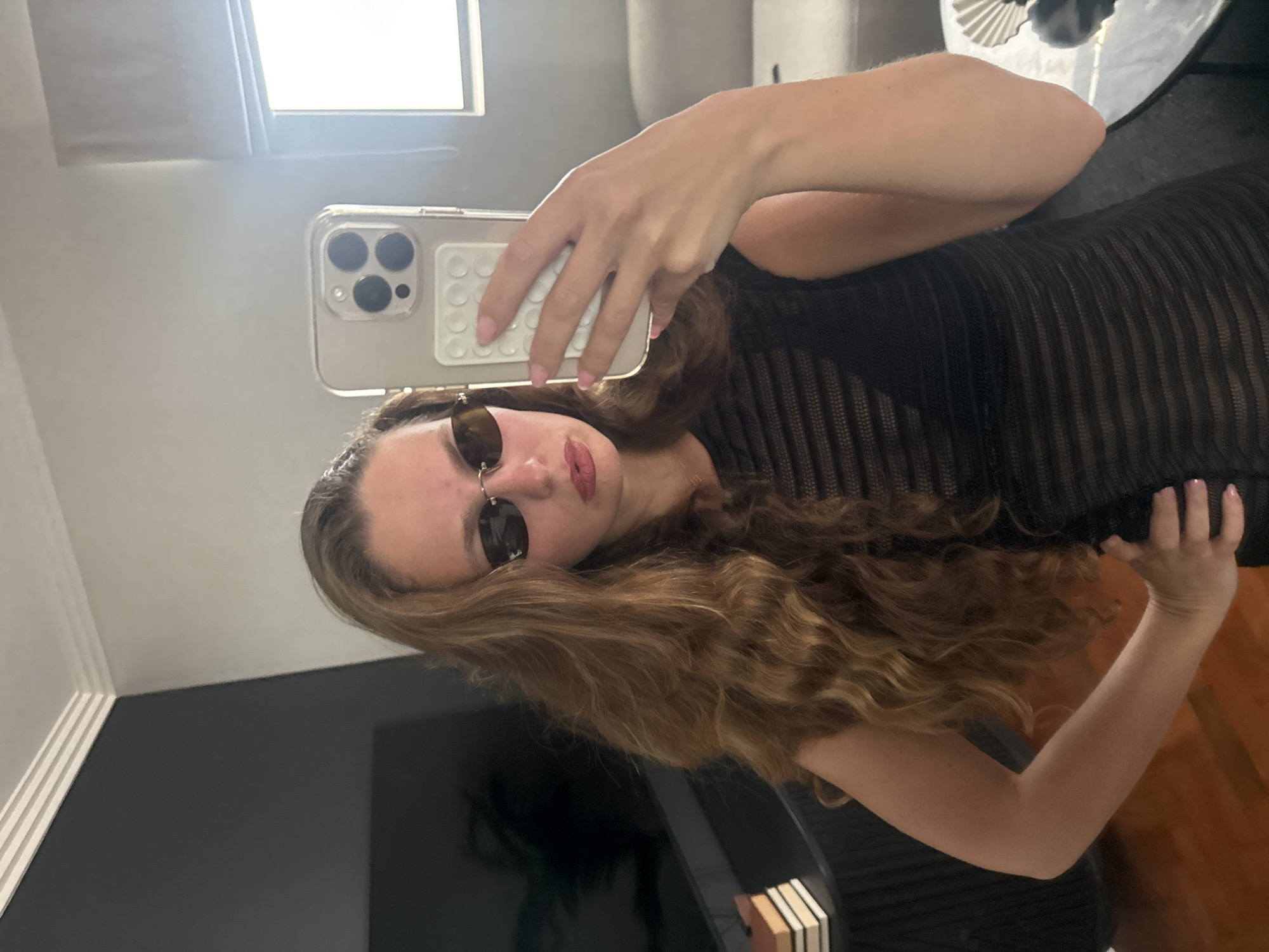 A young woman with long, wavy brown hair takes a selfie with her iPhone in an indoor location. She wears sunglasses and a black, horizontally striped garment. The environment includes a window, wall, and furniture. The photo does not appear to be taken in Cape Town, South Africa.