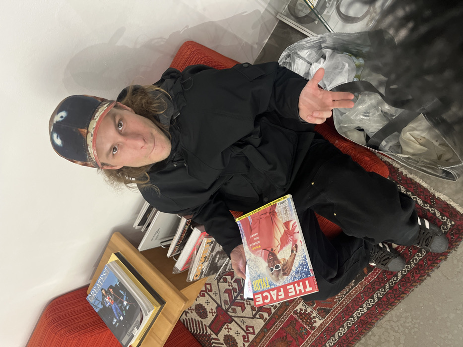 A person in a black jacket and patterned cap sits on an orange cushion. They're holding a magazine titled "The Face" with an image of a person wearing sunglasses on the cover. One hand is raised, forming a finger gun gesture. To the left, there's a stack of books on a wooden shelf, and a white bag is on the right. The person is positioned on a colorful, patterned rug.