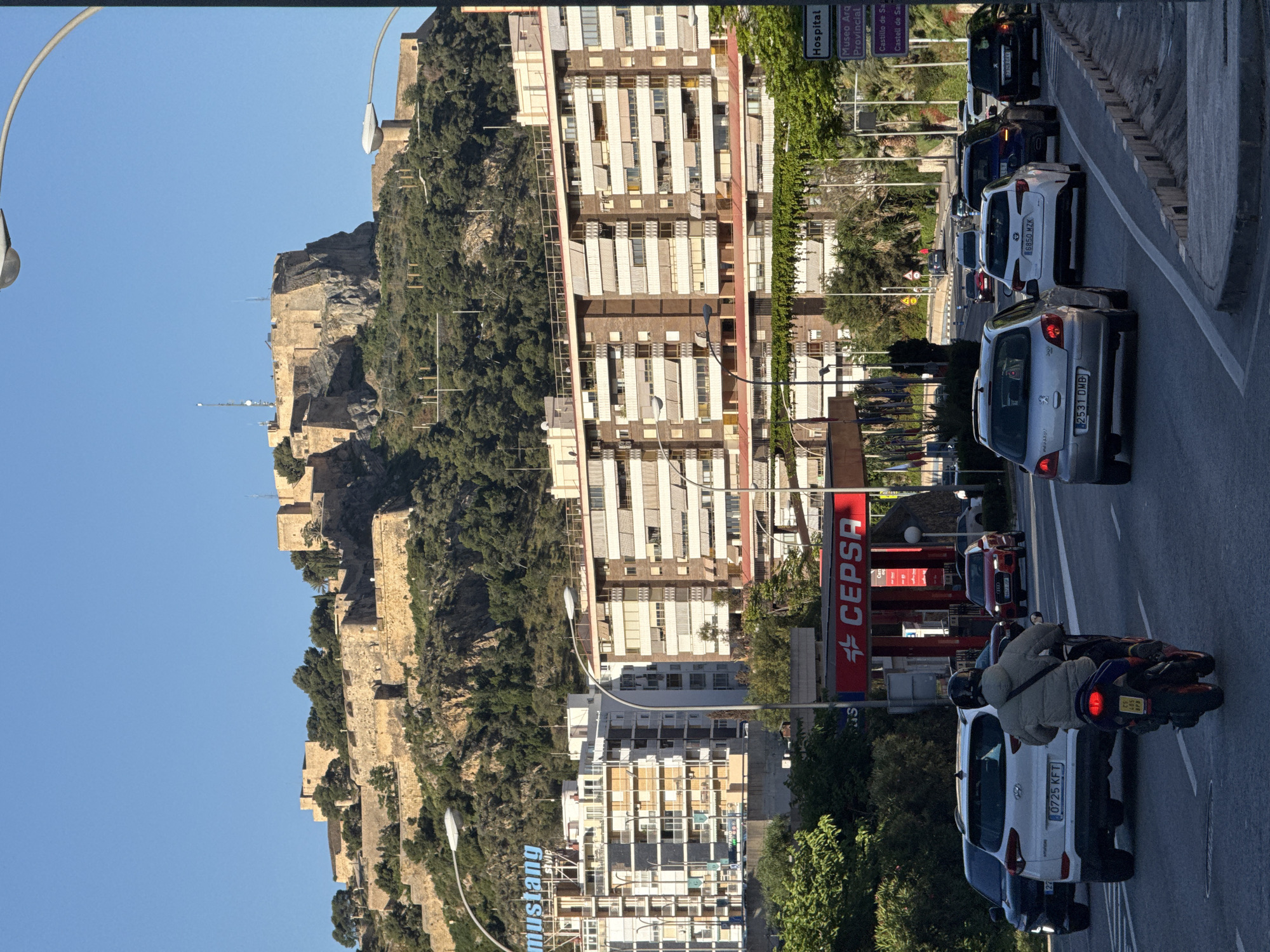 The image captures a street scene in an unknown location, featuring a mix of historical and modern elements. A castle structure, possibly a historical landmark, sits atop a rocky hill covered in trees. Alongside the hill, there are several multi-story buildings with repetitive window patterns, suggesting residential or commercial use.
The foreground shows a busy street with cars and a motorcycle in traffic. Visible signs include a "CEPSA" gas station and signs indicating "Hospital" and "Museo Arq Provincial" (Archaeological Provincial Museum), pointing to the presence of healthcare and cultural institutions in the area. The street includes several streetlights. A "mustang" sign is also visible in the distance.