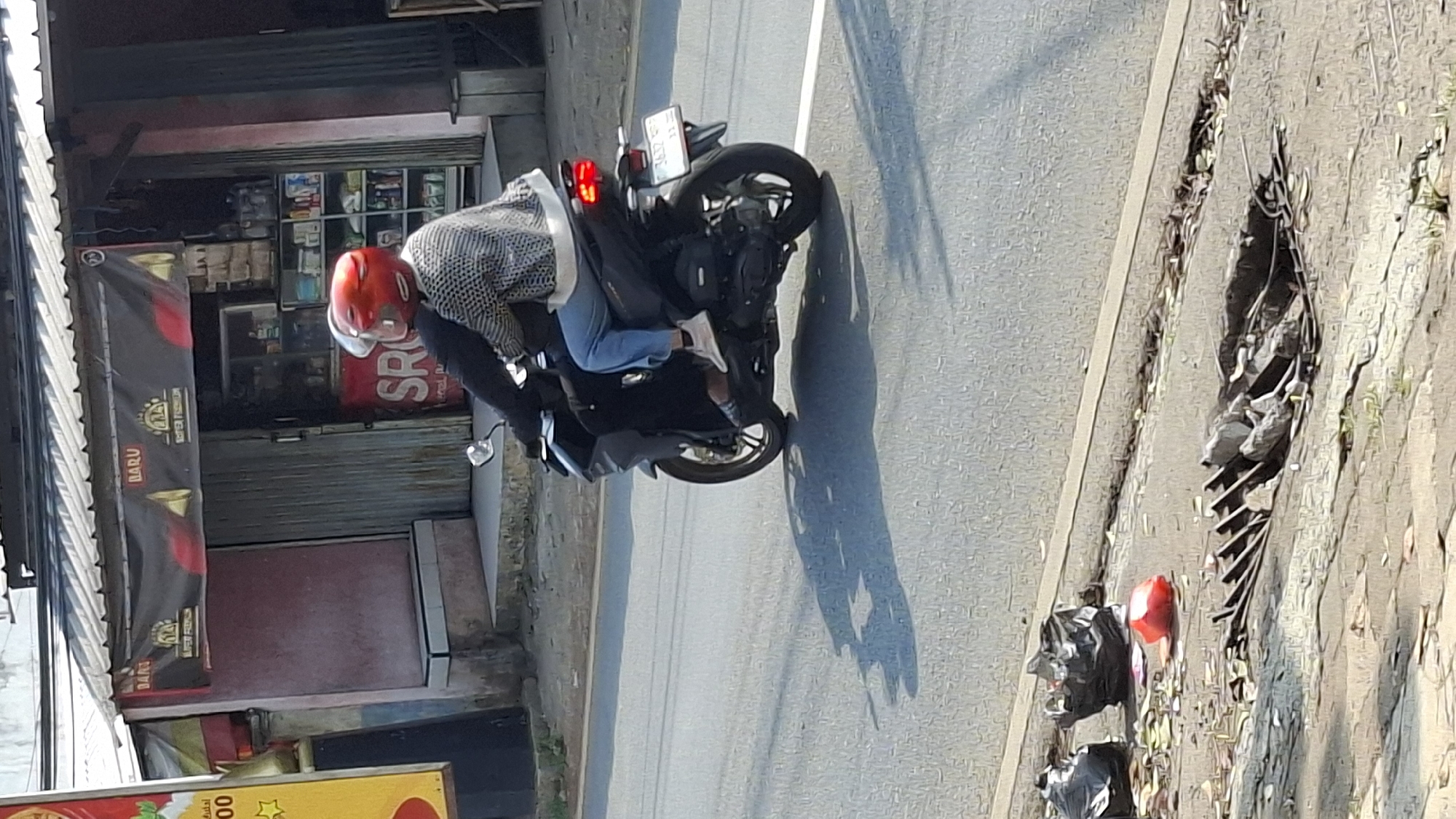 Two individuals, a driver and a passenger, are riding a dark-colored motorcycle on a paved road. The driver is wearing a black jacket and the passenger is wearing a gray and white patterned jacket and blue pants. Both are wearing helmets, one red and one white. The motorcycle has a white license plate with dark lettering. In the background, there is a building with a black awning that displays the word "BARU" and other text. Shelves with various products are visible inside the building. In the foreground, on the side of the road, there are several black trash bags, a red object, and debris including leaves and some metal structures, suggesting a drain or damaged infrastructure. The scene appears to be an outdoor urban or semi-urban environment in Cilaku, Indonesia, indicated by the signage in the background. The lighting suggests it is daytime, possibly late morning or early afternoon, given the shadows.